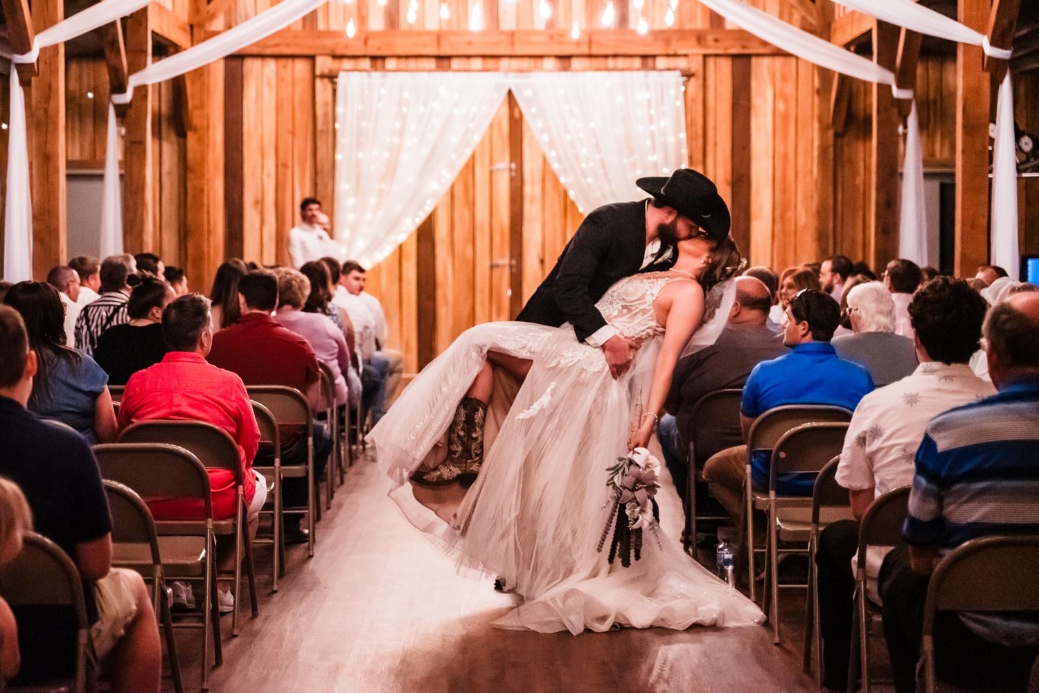 Romantic couple shares their first kiss during a wedding ceremony in a rustic wooden barn filled with guests.