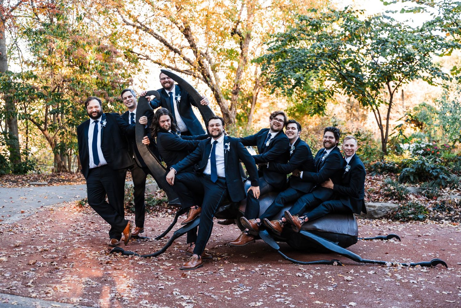 Groomsmen in black suits posing playfully outdoors on a fall day with autumn foliage in the background.