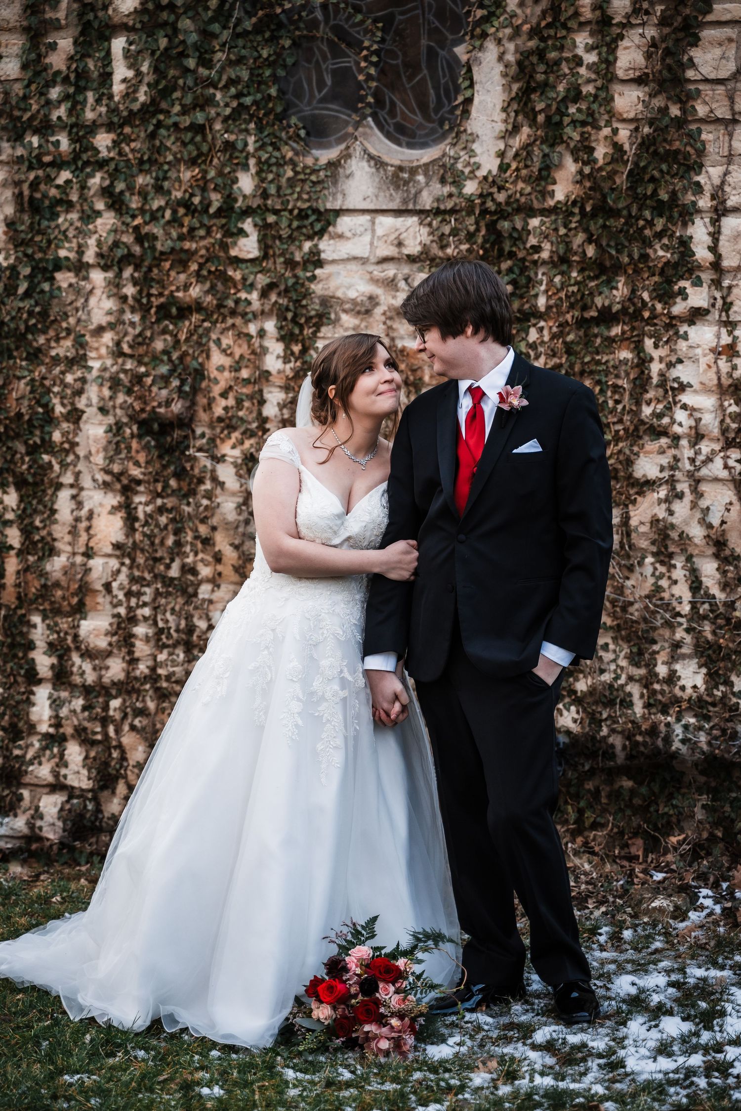 A winter wedding portrait features a couple in formal attire standing against an ivy-covered church wall.