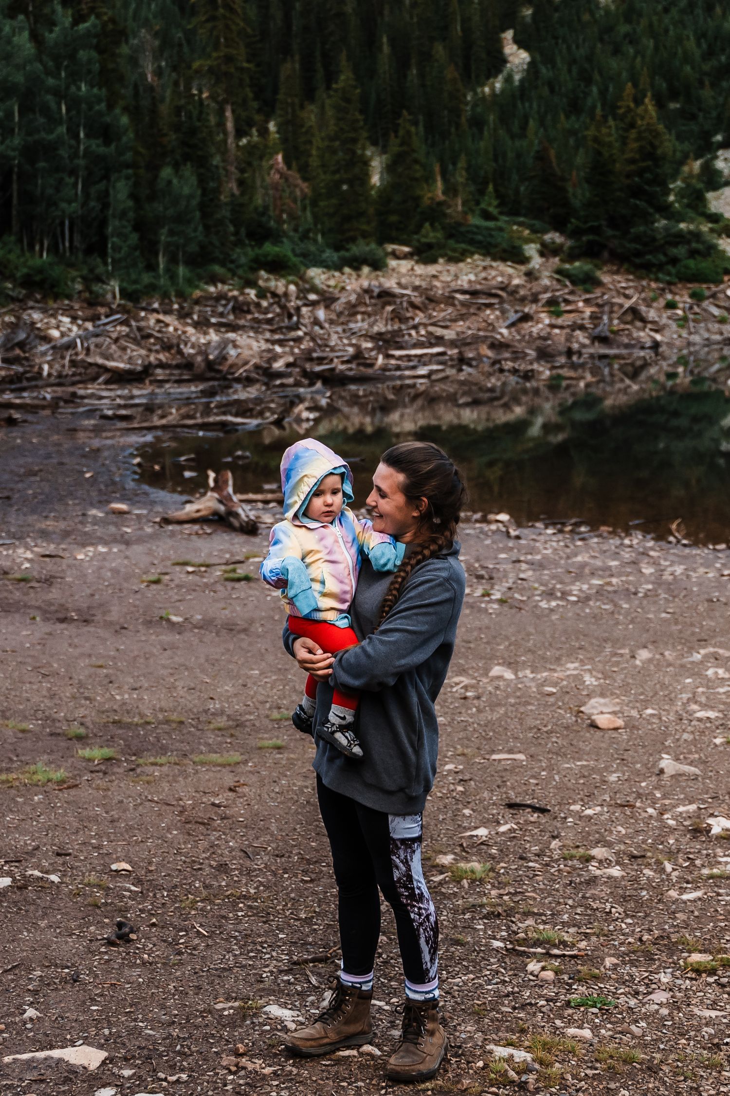 An adult and child stand at the edge of a lake surrounded by pine trees and driftwood in a mountain setting.