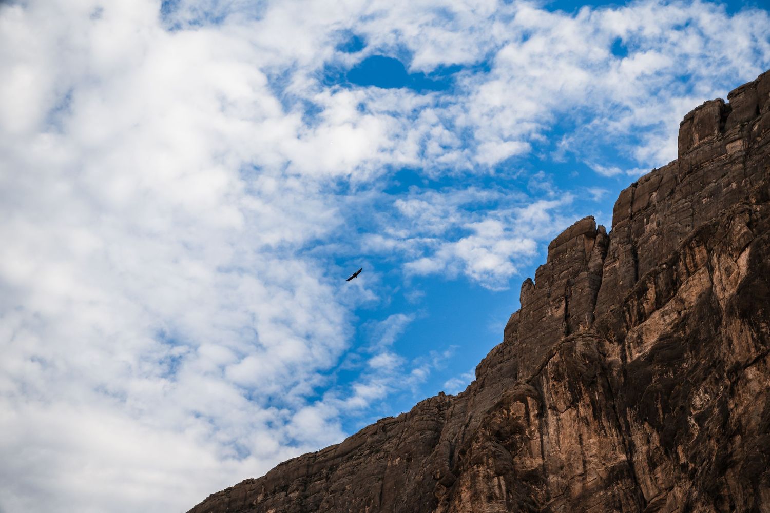 Dramatic rocky cliffs rise against a bright blue sky dotted with white puffy clouds.