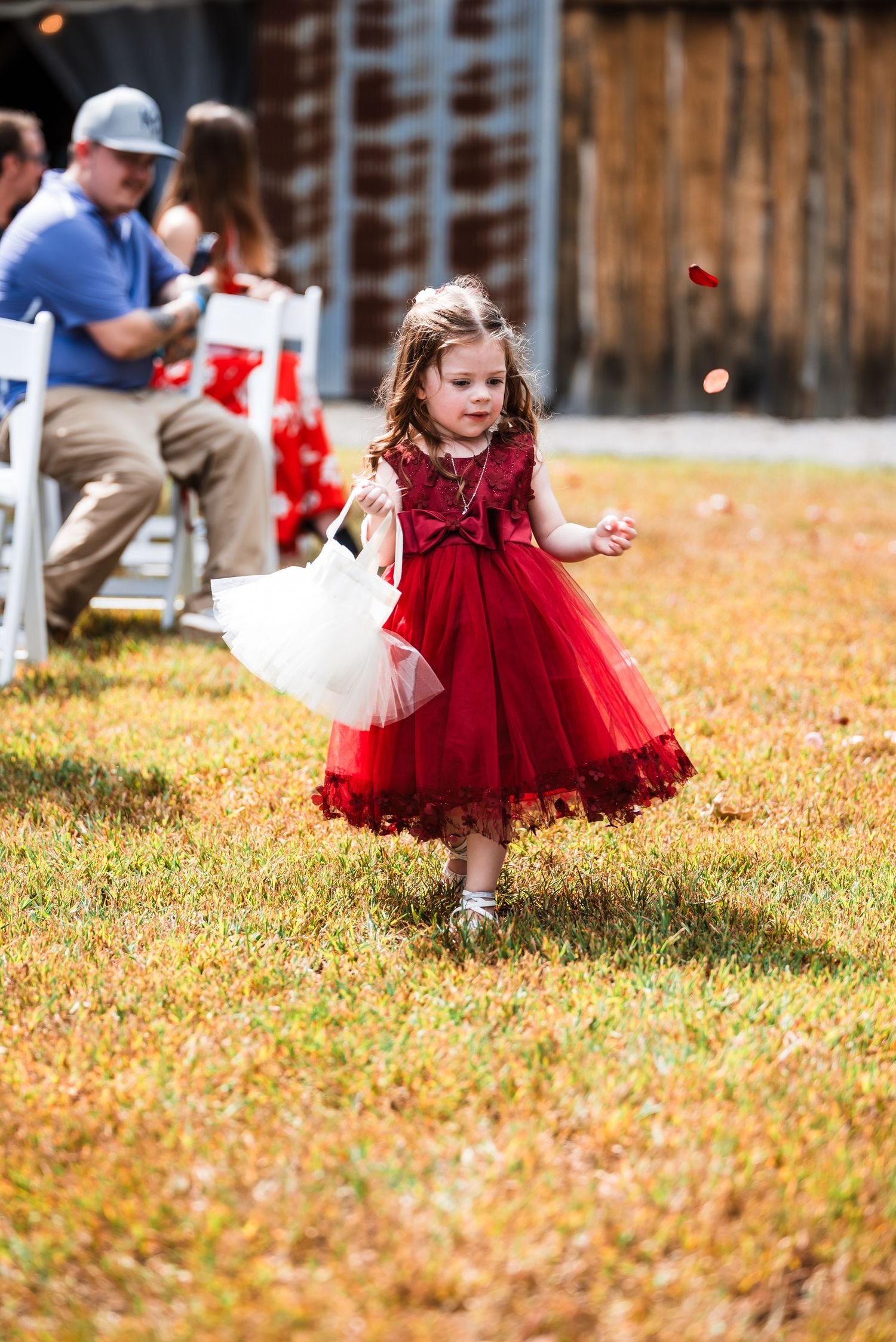 Young flower girl in red dress twirls on autumn grass during wedding festivities.