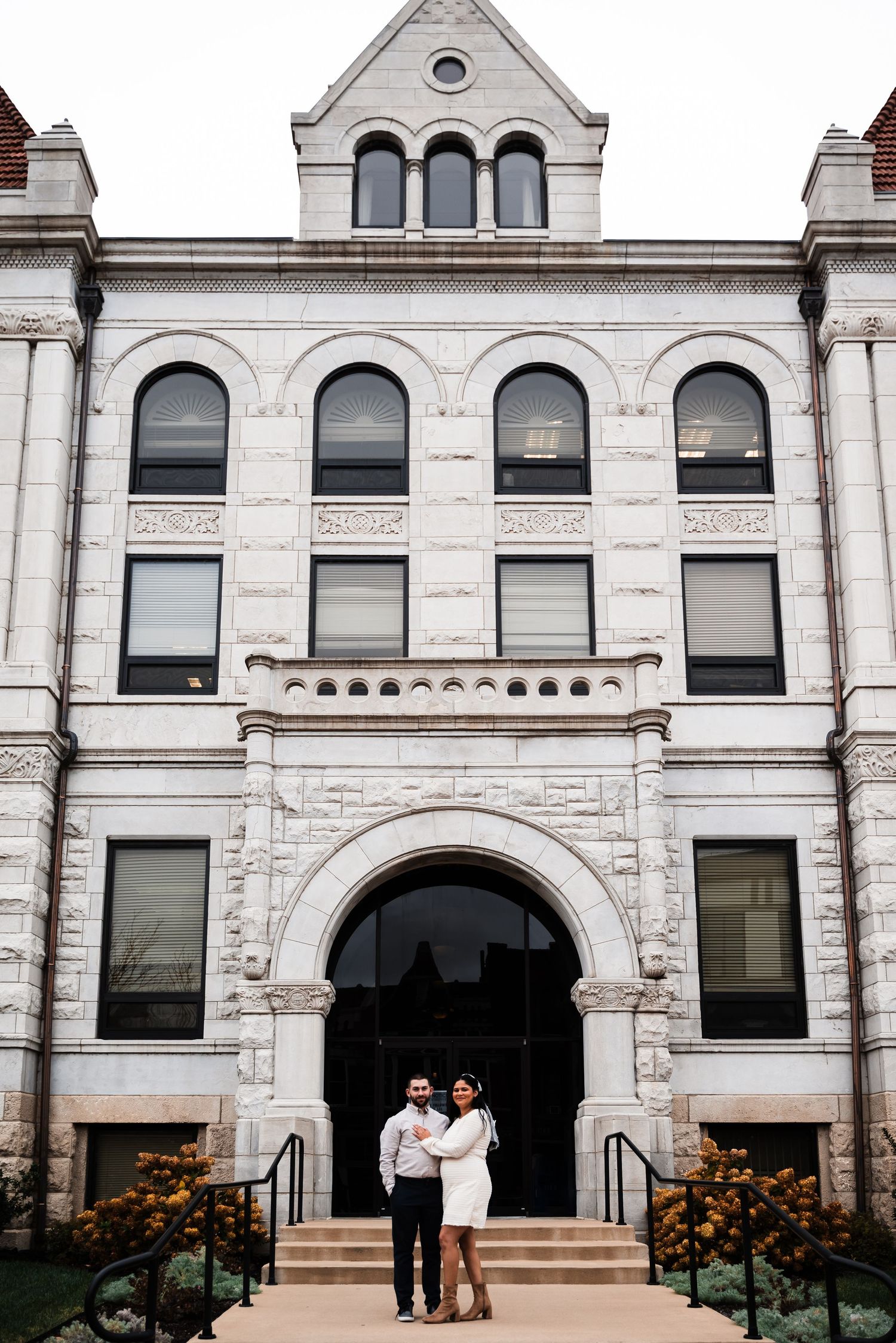 A couple poses in front of a historic white stone building with arched entrance and ornate architectural details.