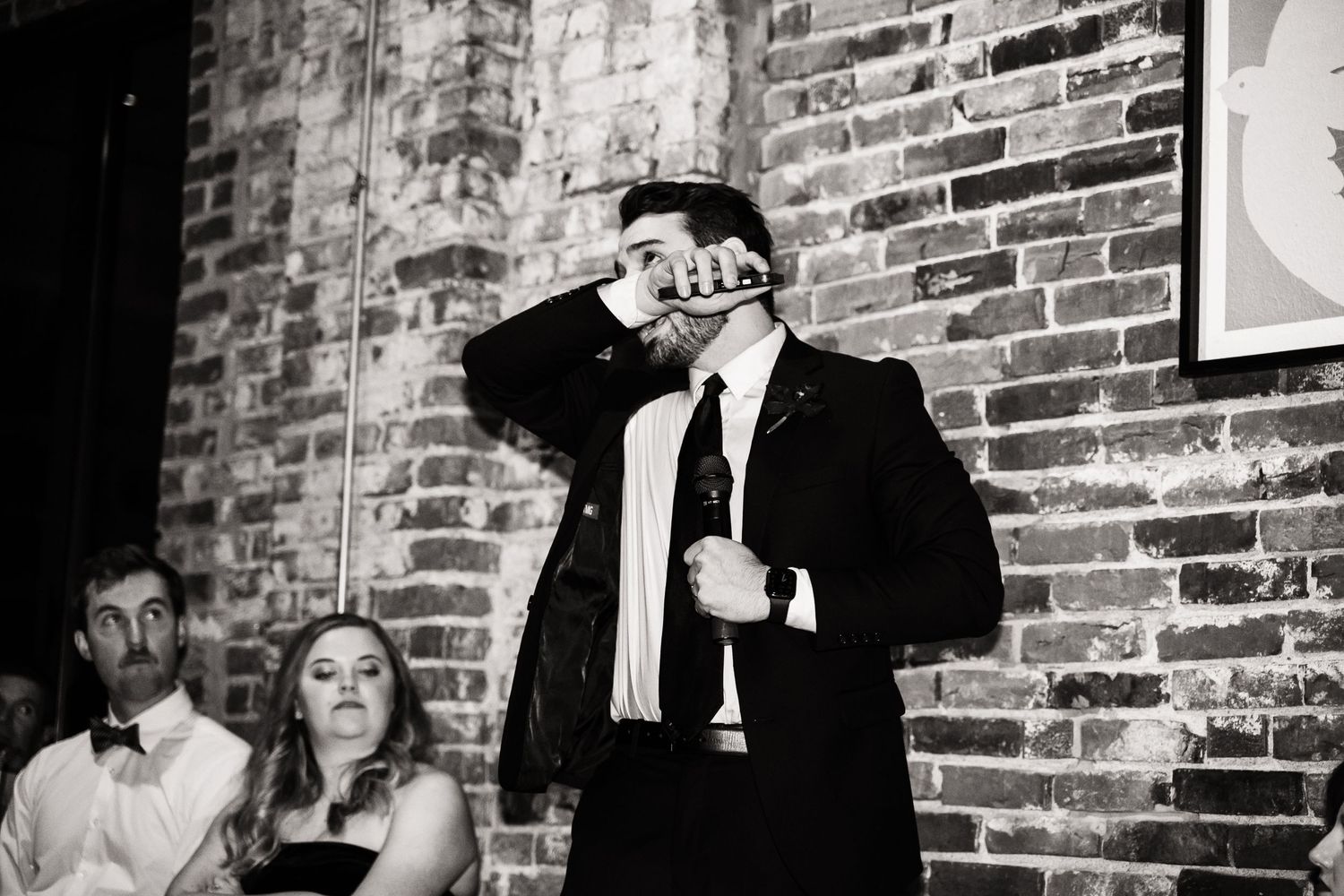 Black and white candid shot of person giving speech at brick-walled wedding reception venue.