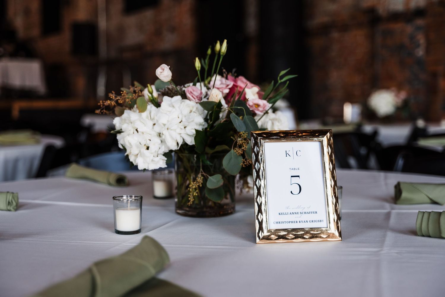An elegant table setting with pink and white floral centerpiece, glowing votive candle, and gold-framed table number 5 card.