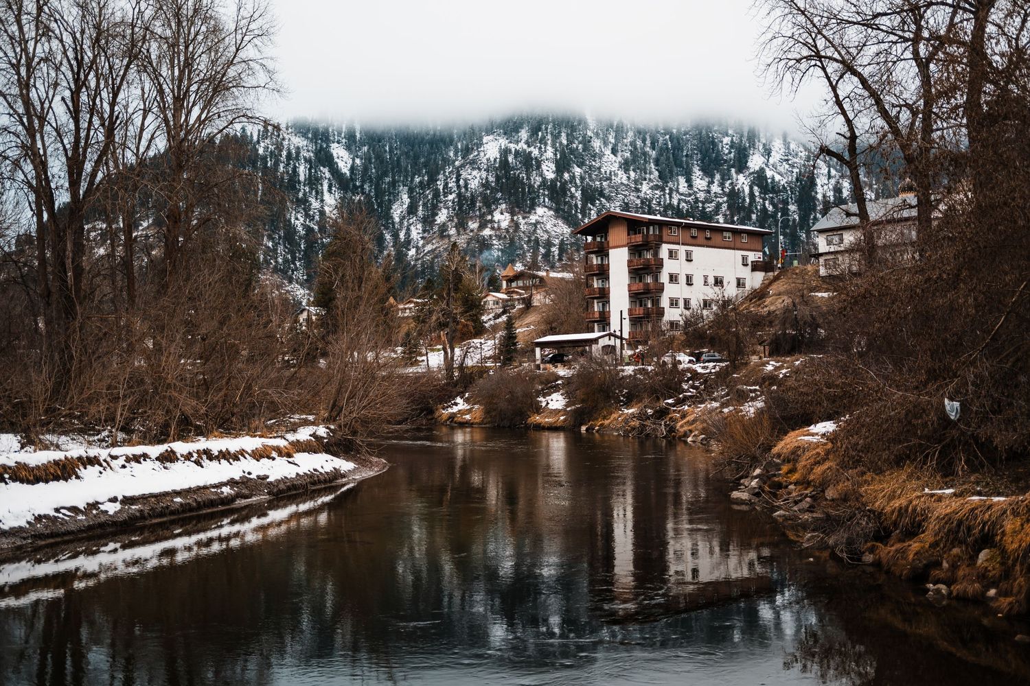 Rustic white building reflects in calm river waters surrounded by snowy winter landscape and bare trees in mountains.
