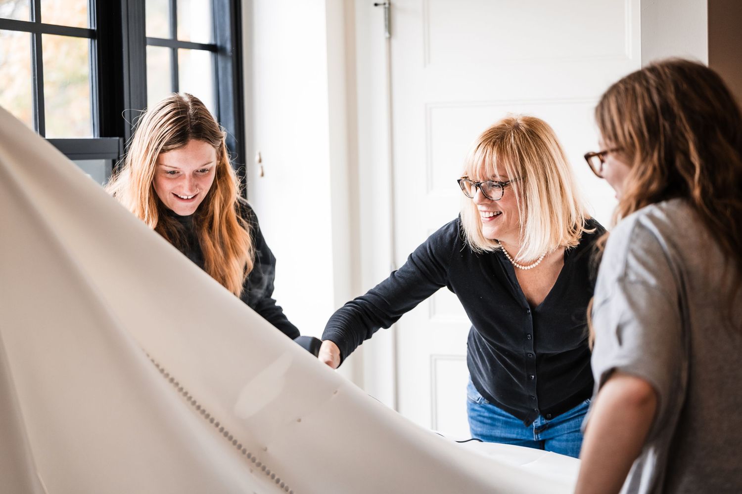 Women examining and touching white fabric in a bright room with large windows.