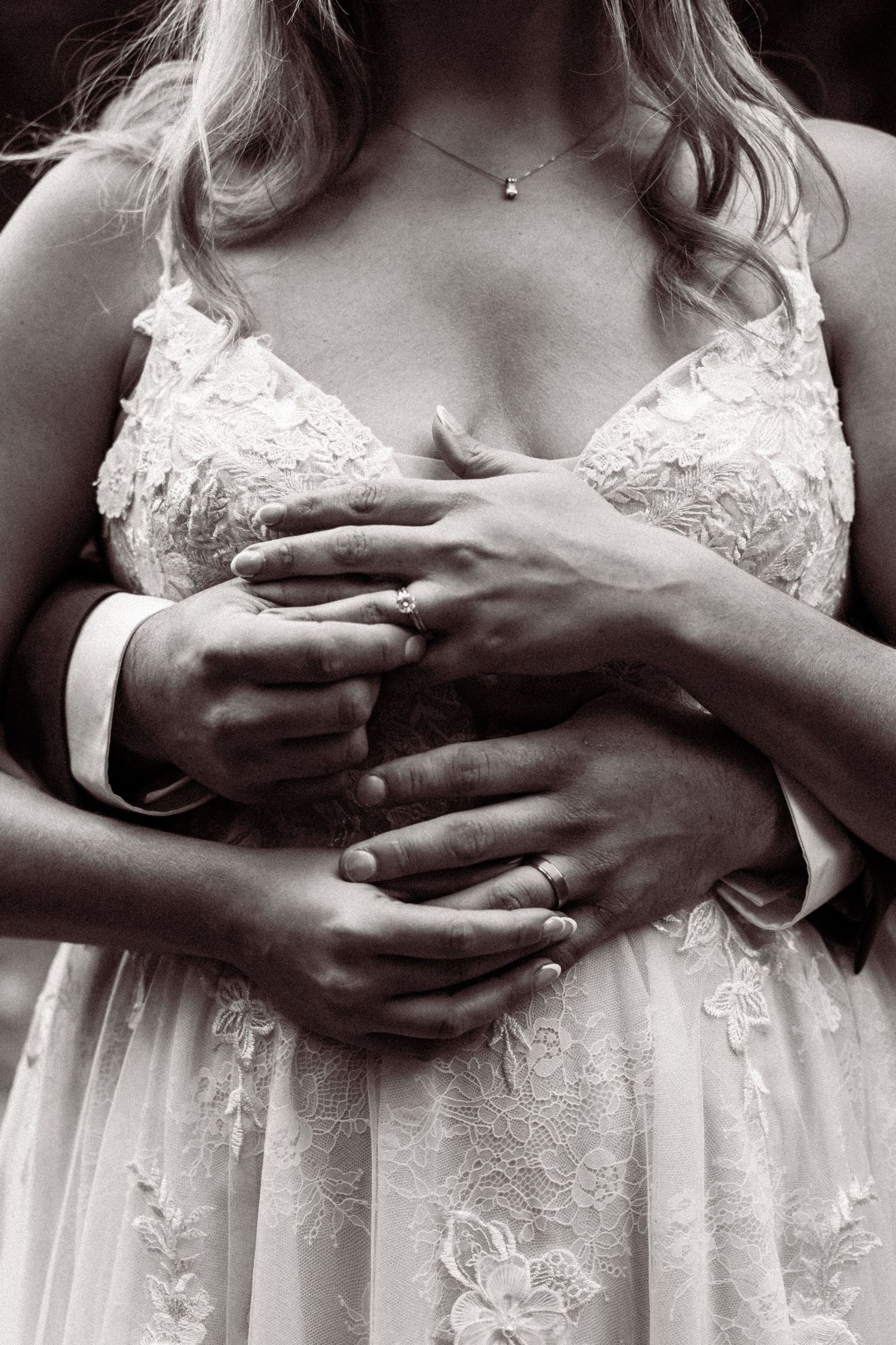 Black and white close-up of hands embracing a torso wearing a delicate lace wedding dress with floral details.