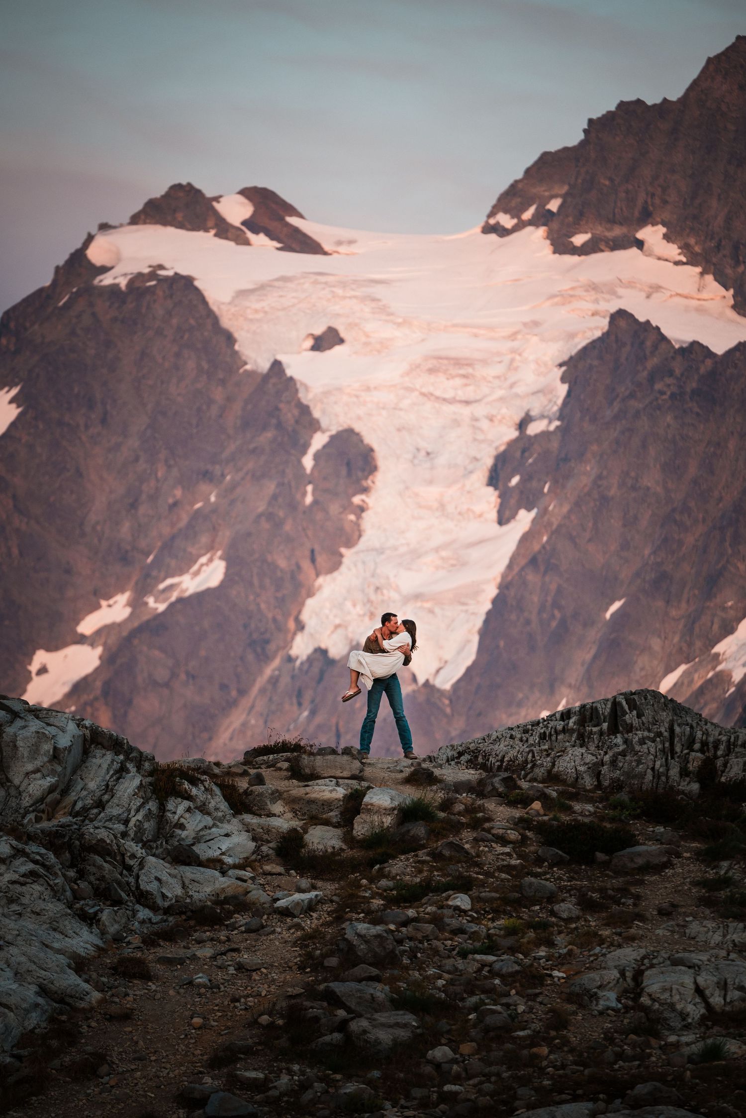 A silhouetted figure stands on a rocky ridge against a dramatic mountain backdrop with snow-capped peaks at sunset.
