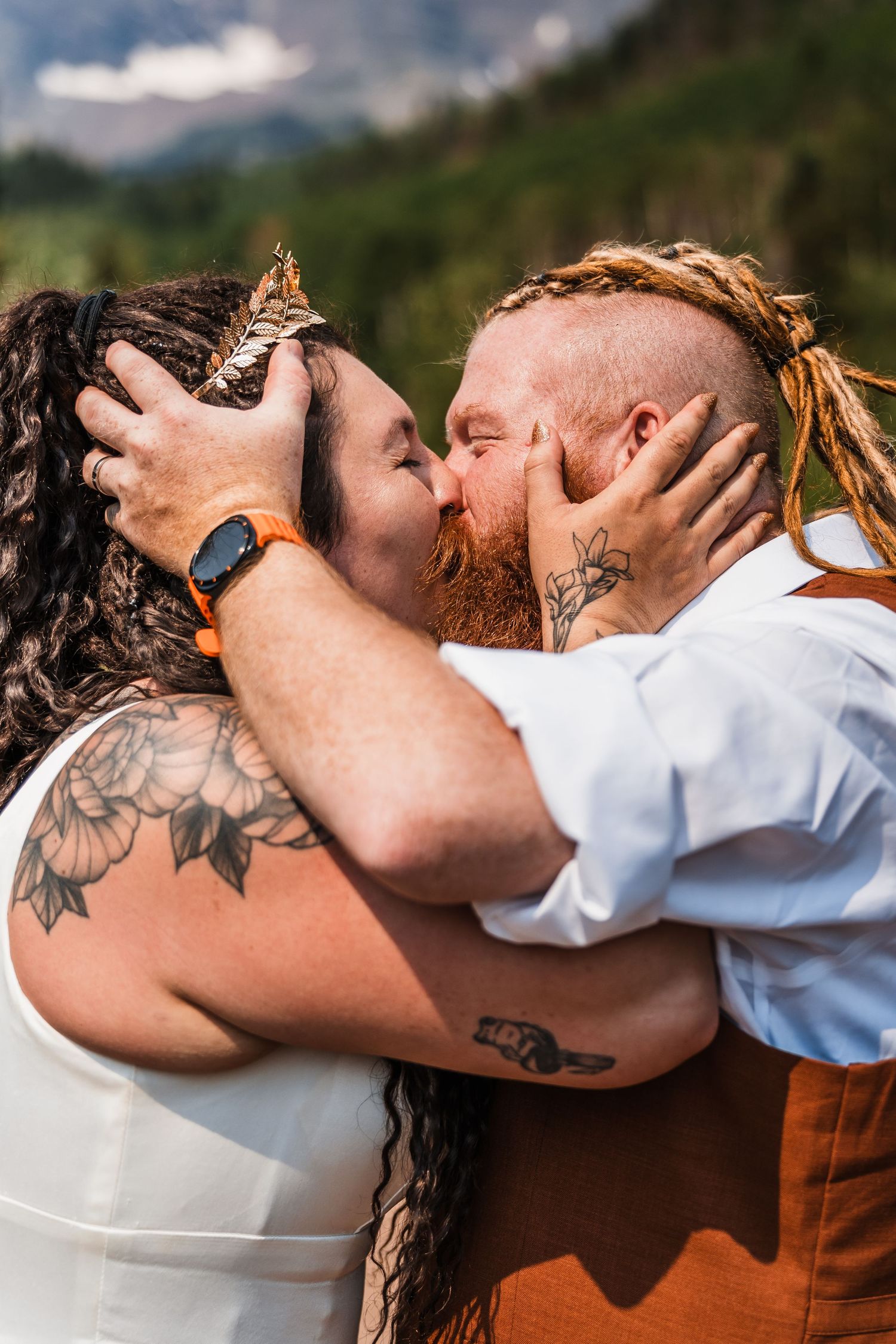 Tattooed couple sharing passionate first kiss during outdoor mountain elopement, bride wearing gold leaf crown.