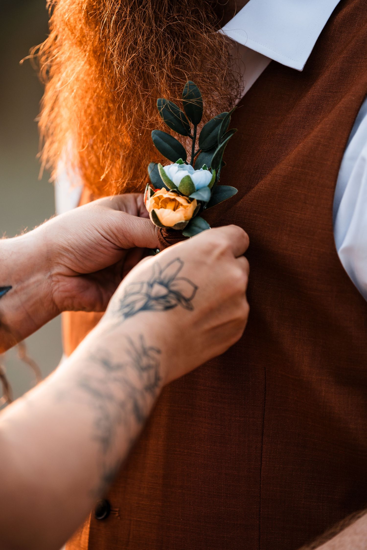 Tattooed hands pinning a small floral boutonniere onto a groom's brown wedding vest.