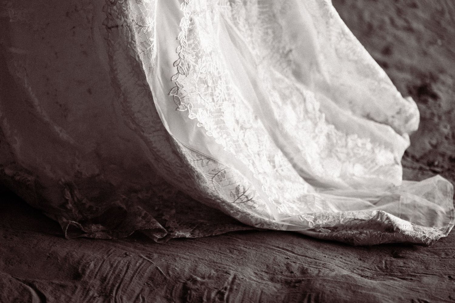 Close-up black and white detail of intricate lace wedding gown train resting on textured rock.