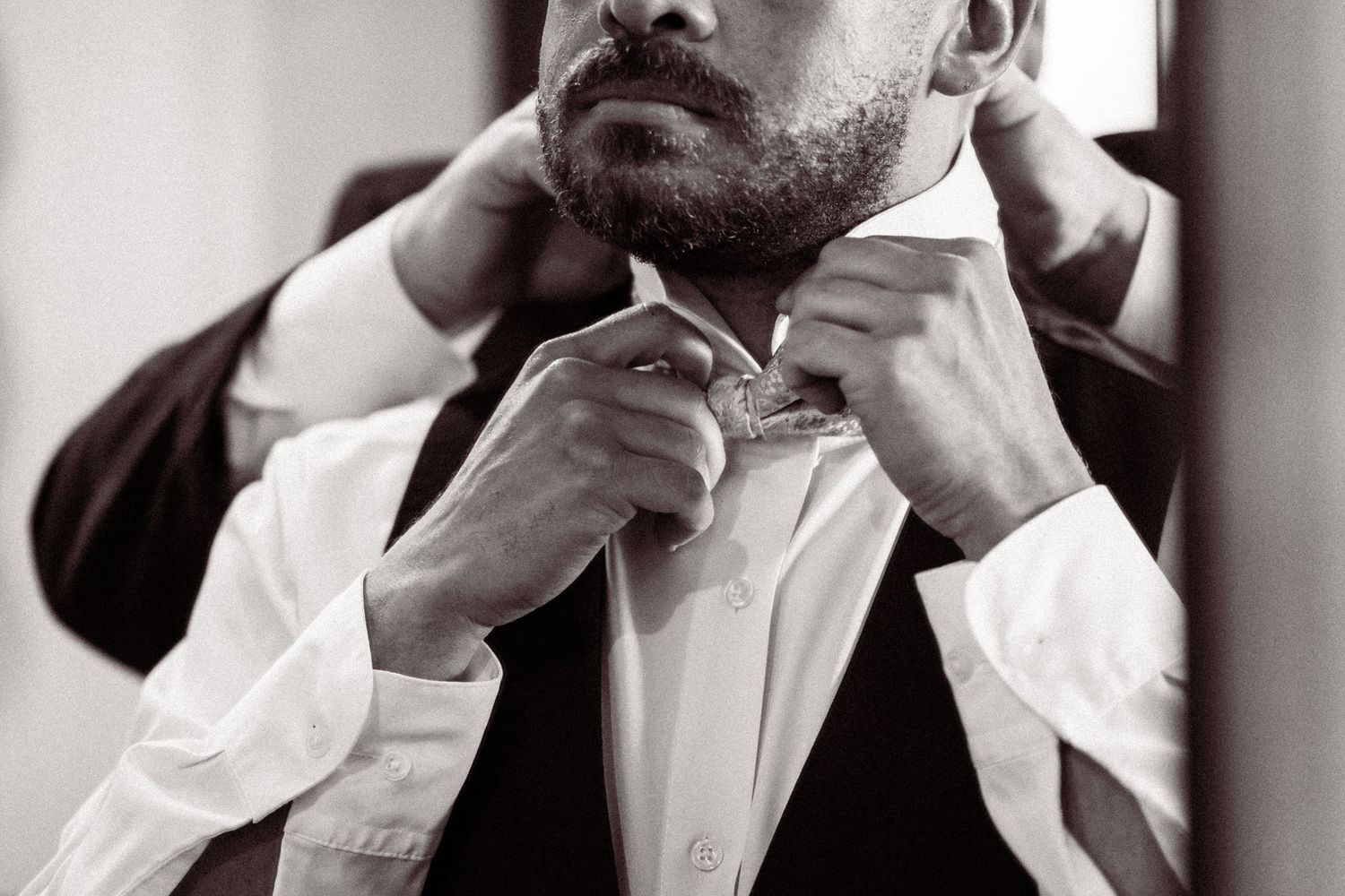 Groom adjusting his bow tie in mirror while getting ready for wedding ceremony in black and white photo.