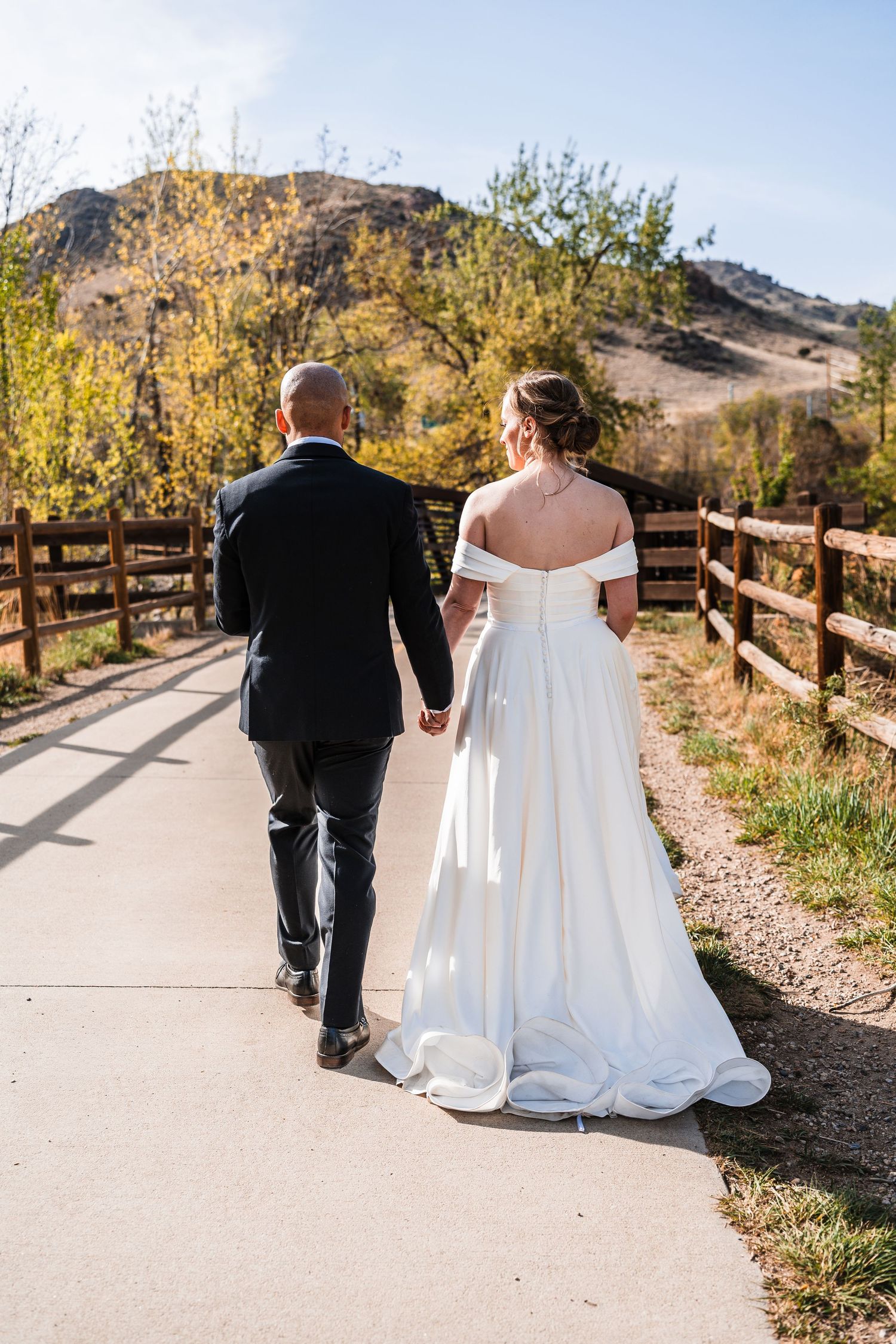 Bride and groom walking hand-in-hand along a scenic mountain path, bride in off-shoulder white gown.