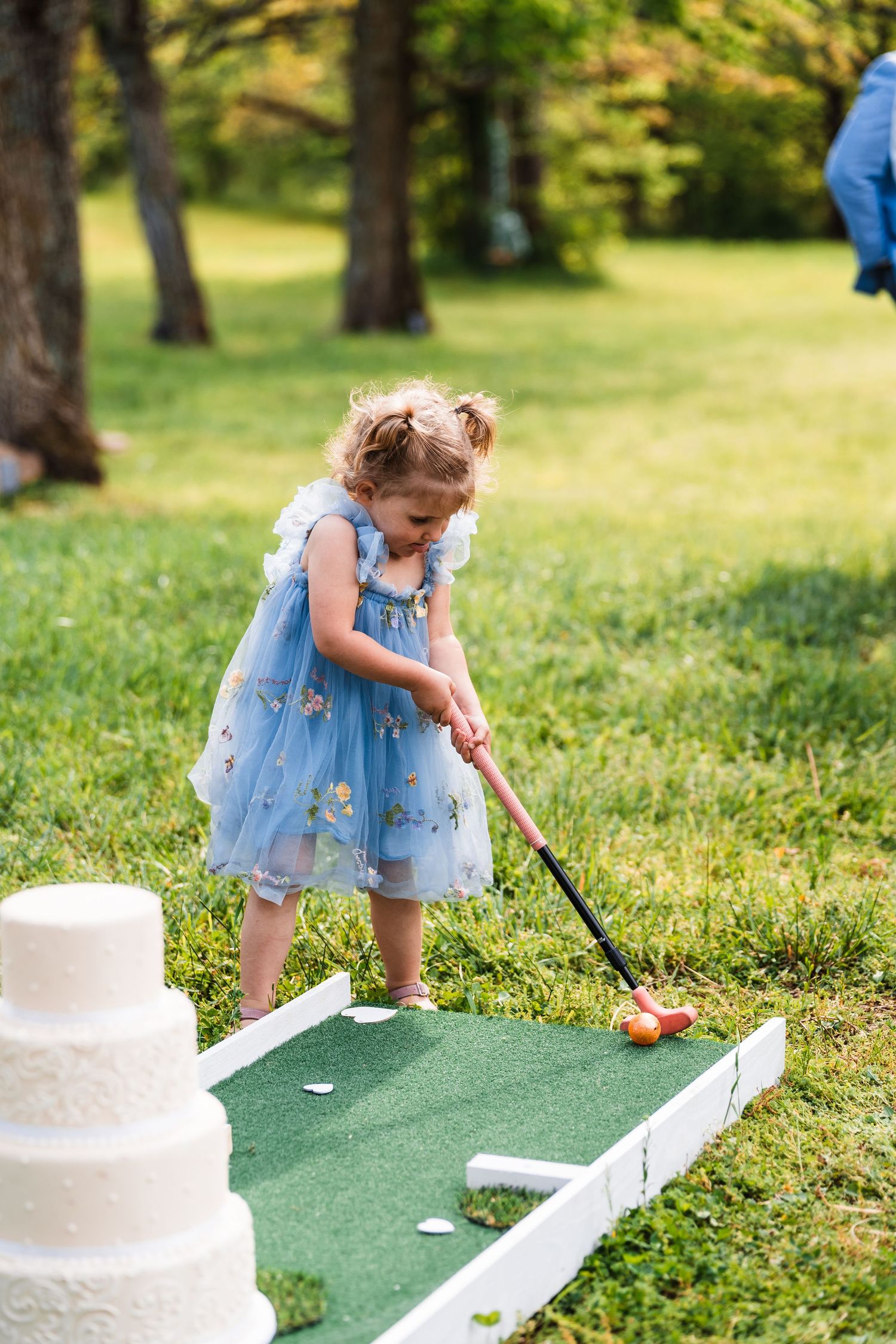 Young flower girl in blue dress playing mini golf near wedding cake at outdoor reception.