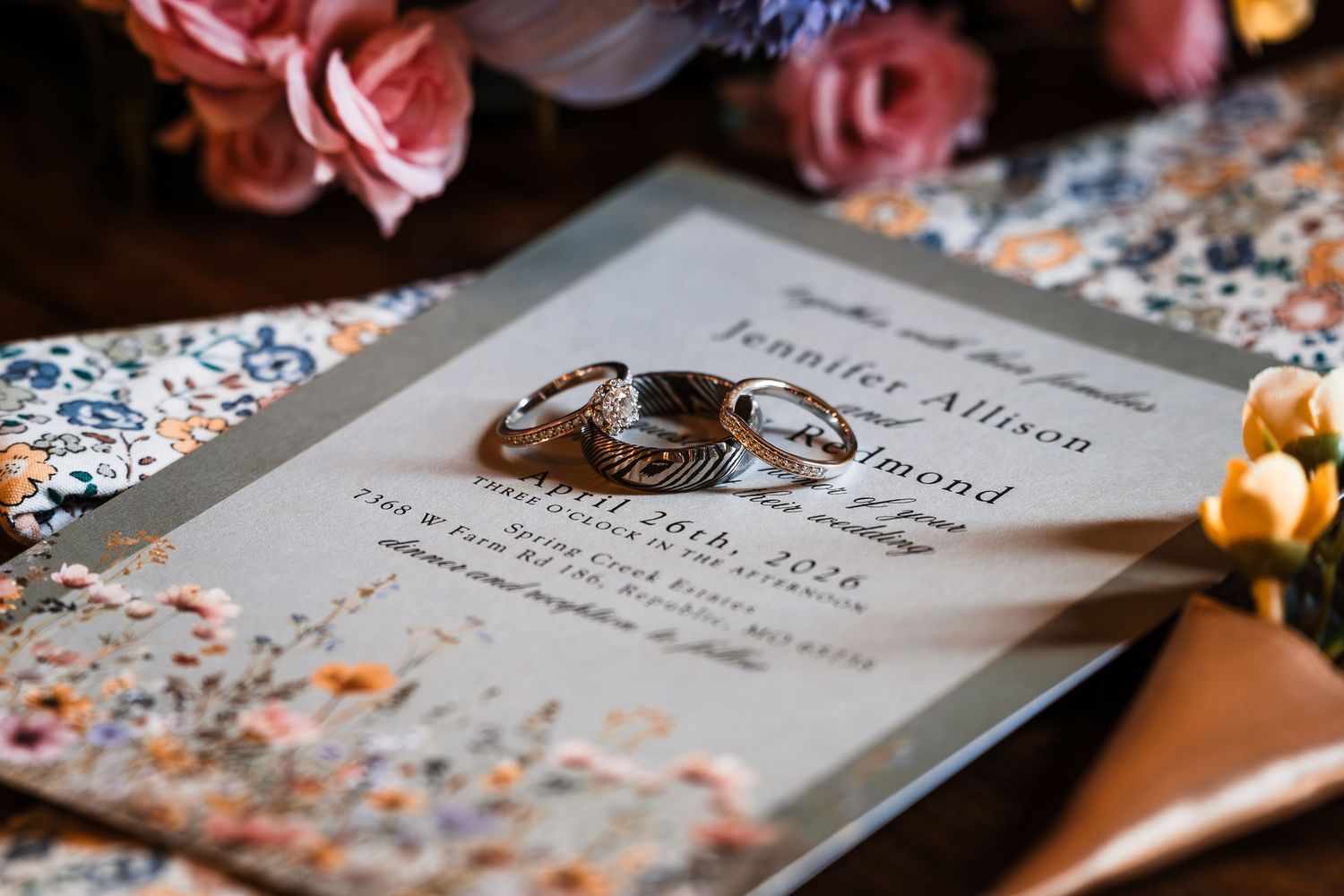 Wedding rings resting on an elegant floral invitation for Jennifer Allison, surrounded by pink roses and decorative paper.