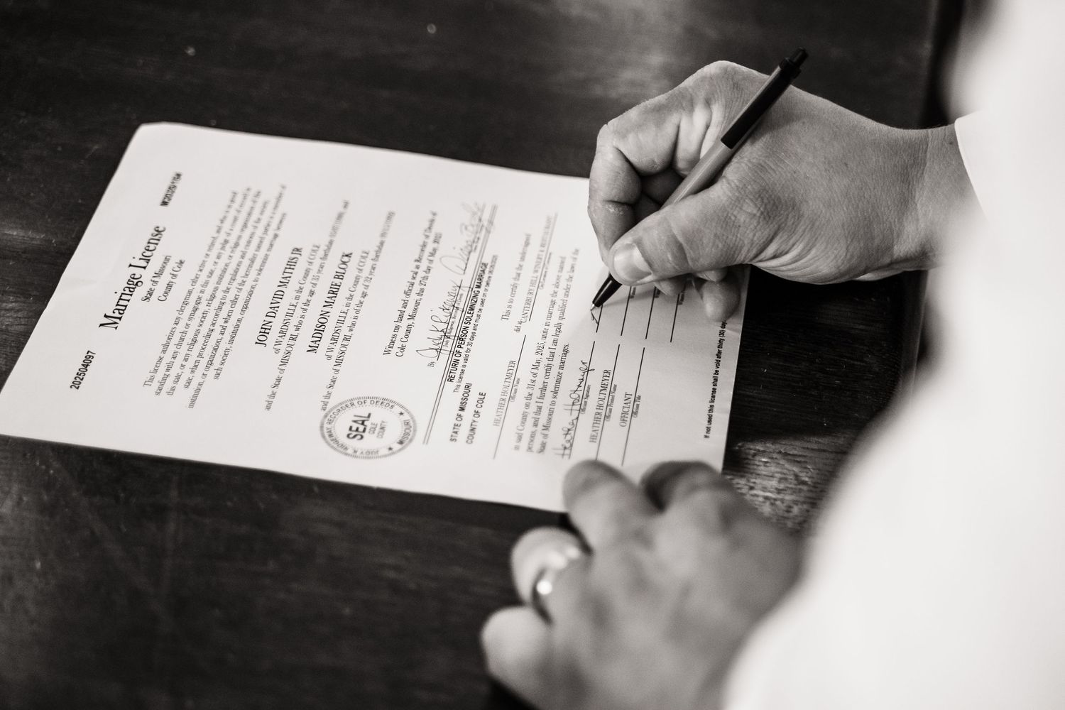 Black and white image showing hands signing an official document or certificate on a formal paper.