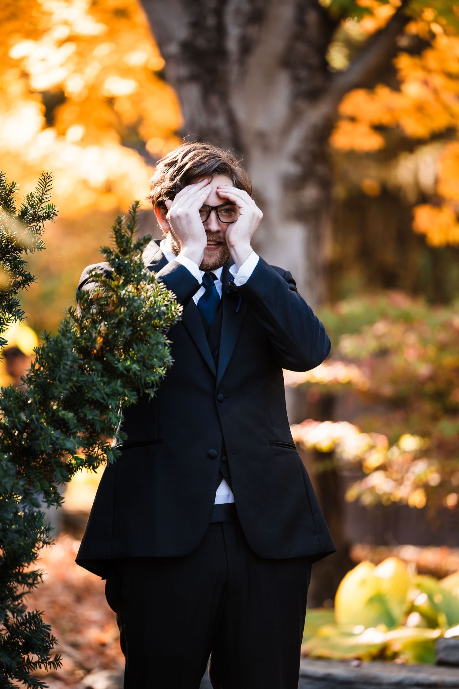 Person in formal attire poses playfully among fall foliage with hands covering face.