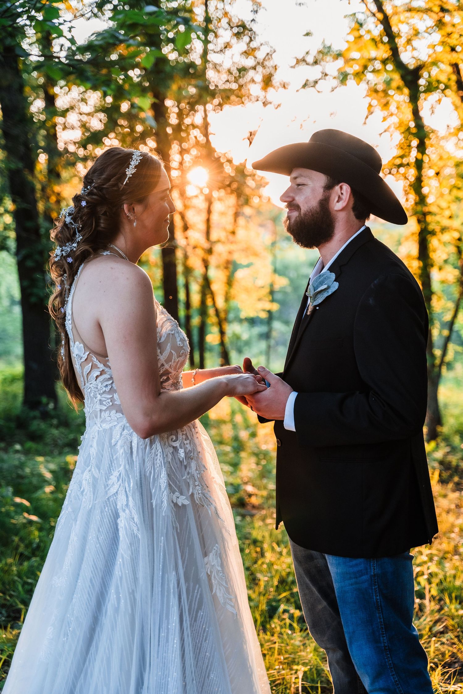 A couple in wedding attire exchange rings during a sunset ceremony in a wooded setting.