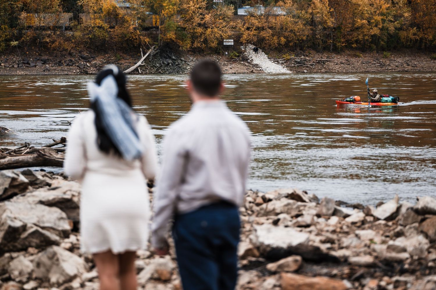 Two people standing on rocky shore watching kayakers paddle by on autumn river.
