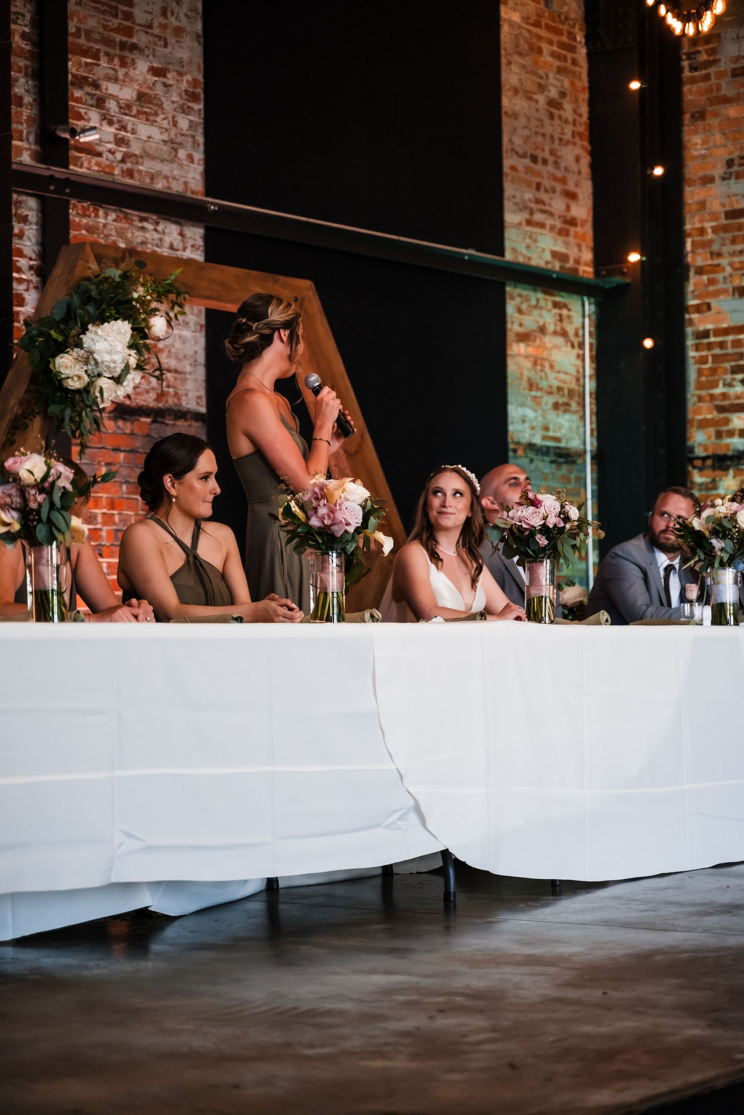 A wedding party sits at a head table with white linens and floral centerpieces against exposed brick walls.