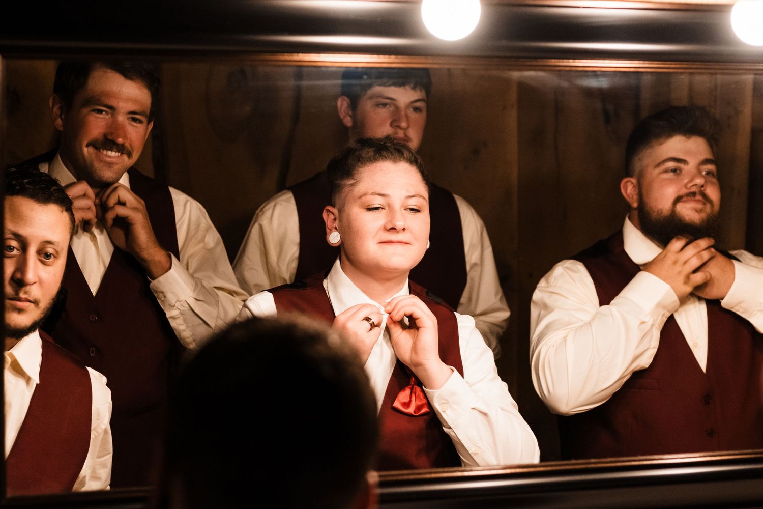 Groomsmen in burgundy vests pose for a wedding party photo in warm lighting.