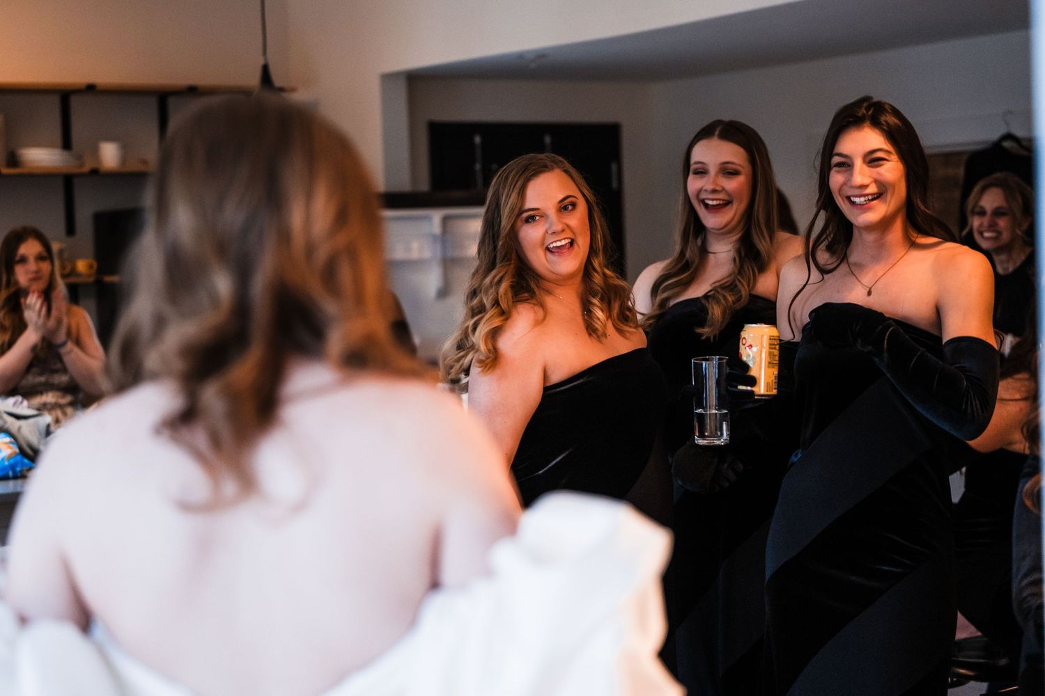 Group of bridesmaids in black dresses share celebratory drinks and laughter.