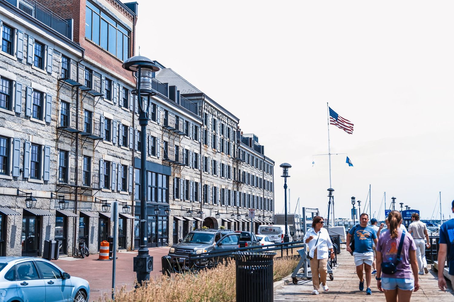 Pedestrians walk along the waterfront boardwalk in Portland, Maine with historic brick buildings and an American flag visible.