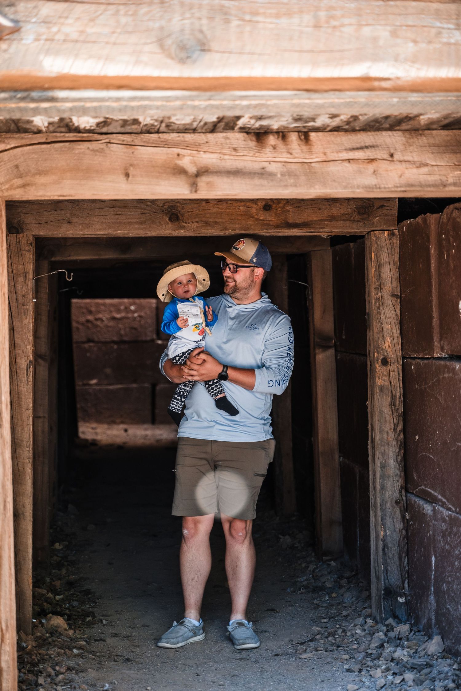 Person standing in a rustic wooden tunnel entrance holding a small child, both dressed casually for exploring.