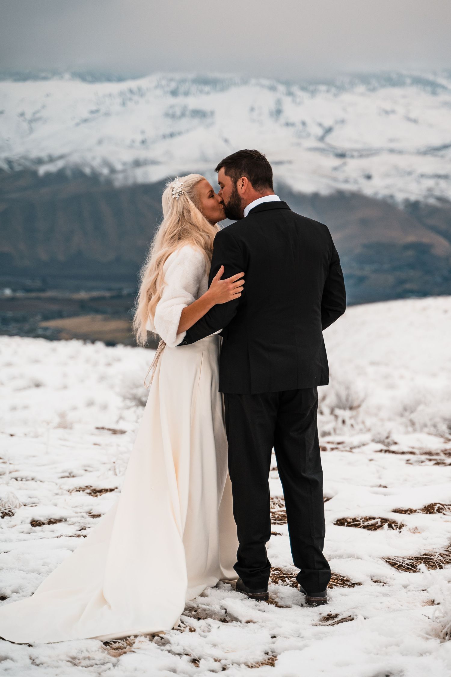 Winter wedding couple embracing on snowy mountain peak with dramatic landscape.