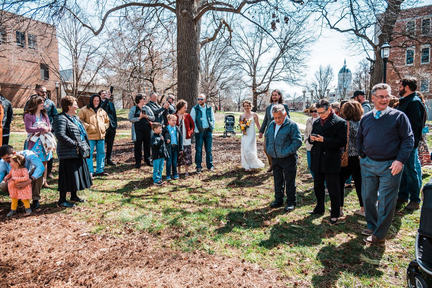 Gathered crowd watching an outdoor ceremony beneath trees on a spring afternoon.
