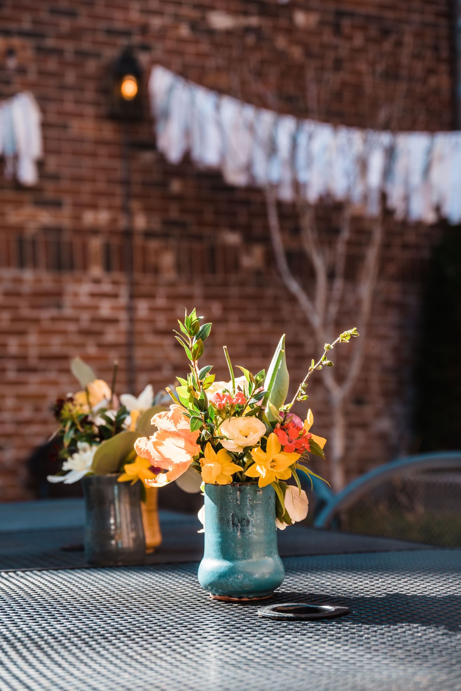 Blue vase with spring flowers on checkered tablecloth against brick wall with white banners.