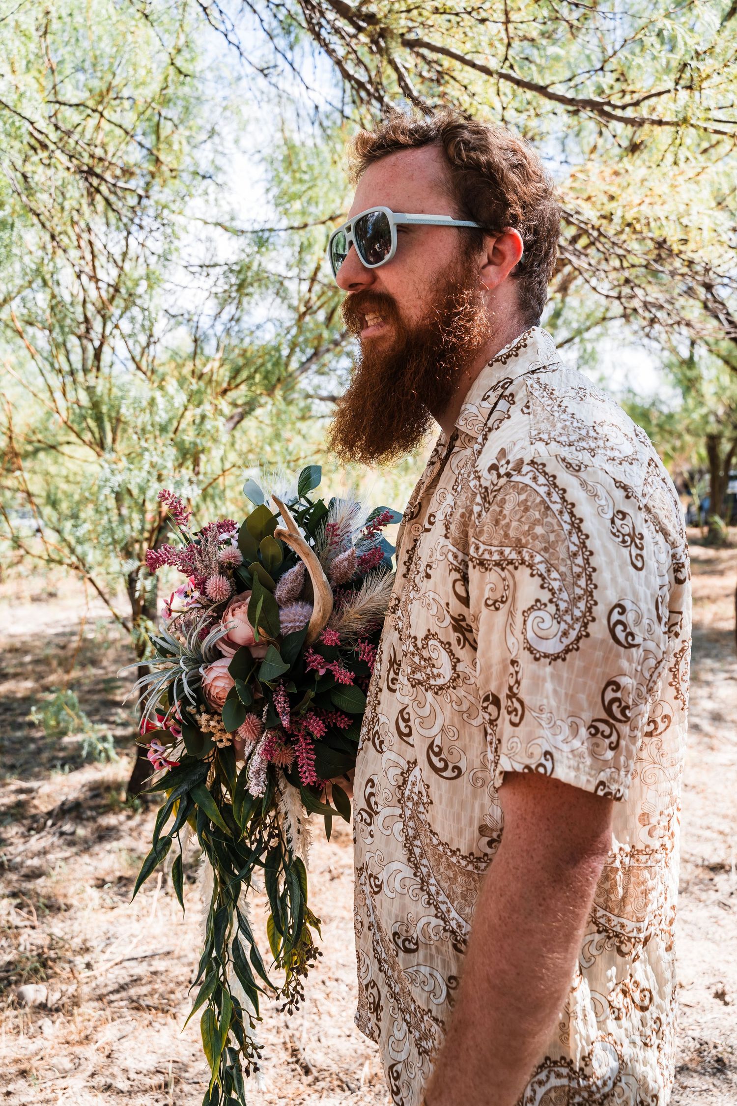 A bearded person in sunglasses and a patterned shirt holds a beautiful bouquet of flowers in a desert setting.