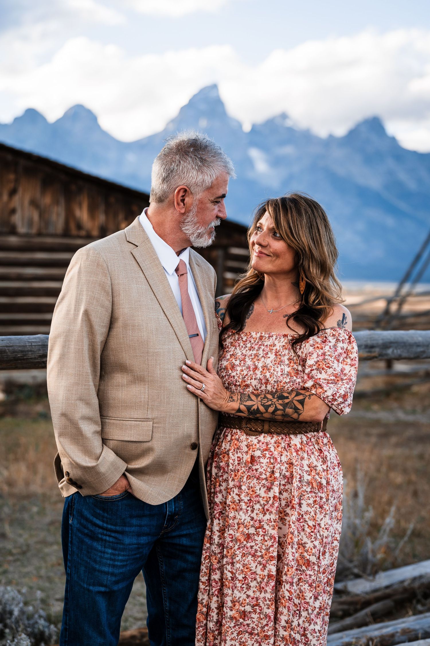 A couple shares a romantic moment against a backdrop of majestic mountains and rustic wooden fencing.