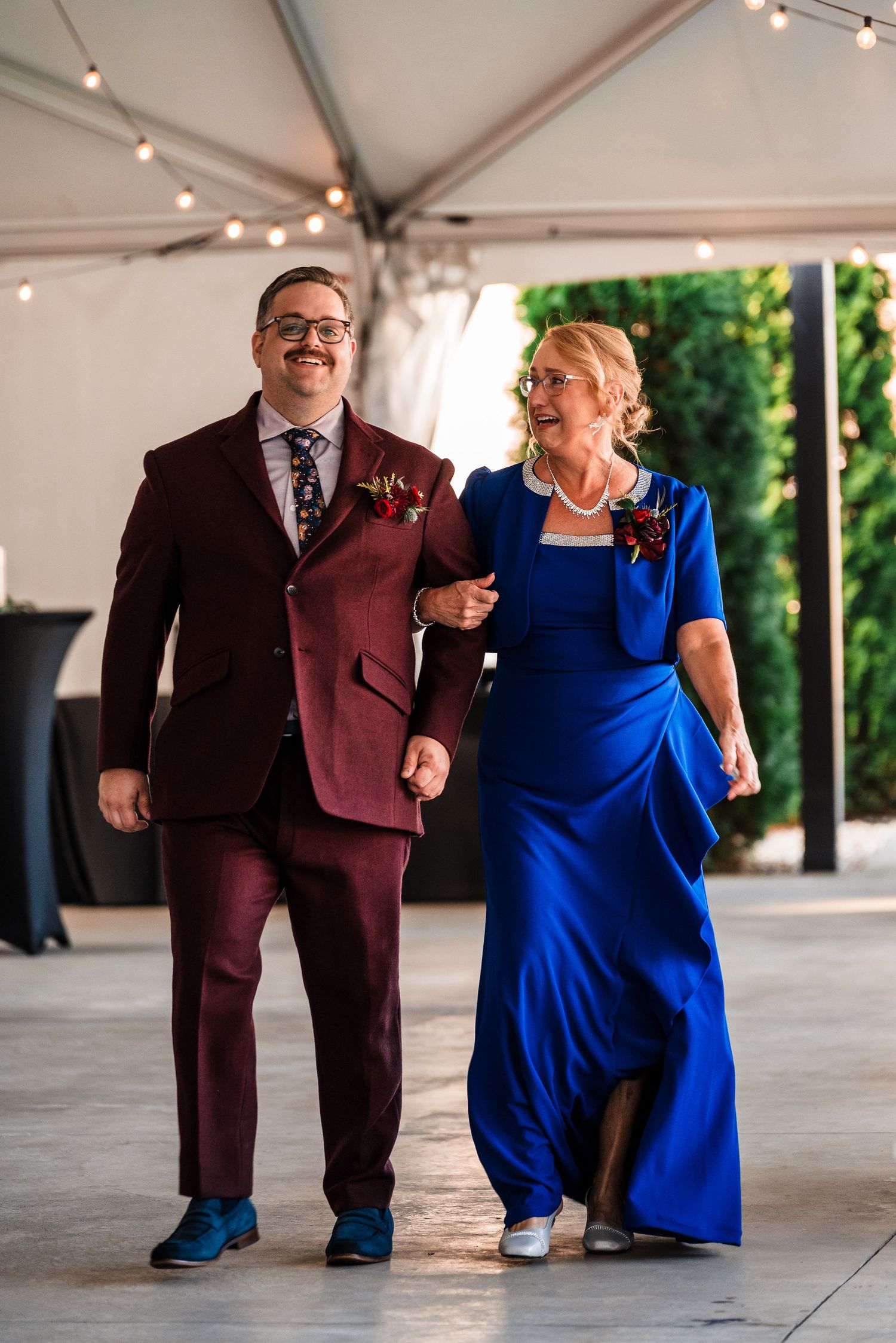 Wedding party members in burgundy suits and royal blue dress walking under a white tent.