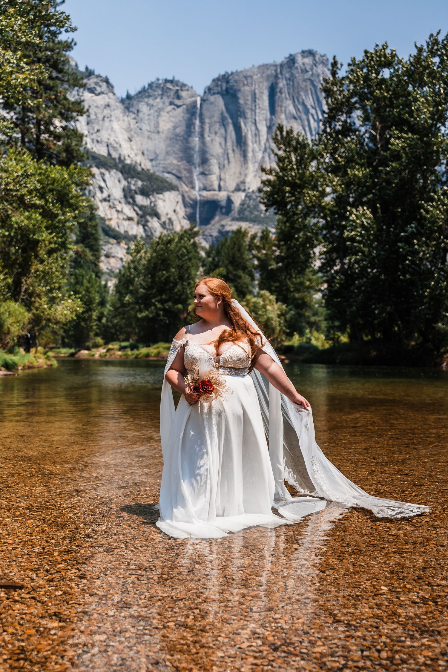 A bride in a flowing white dress stands in shallow water with Yosemite Falls and granite cliffs towering in the background.