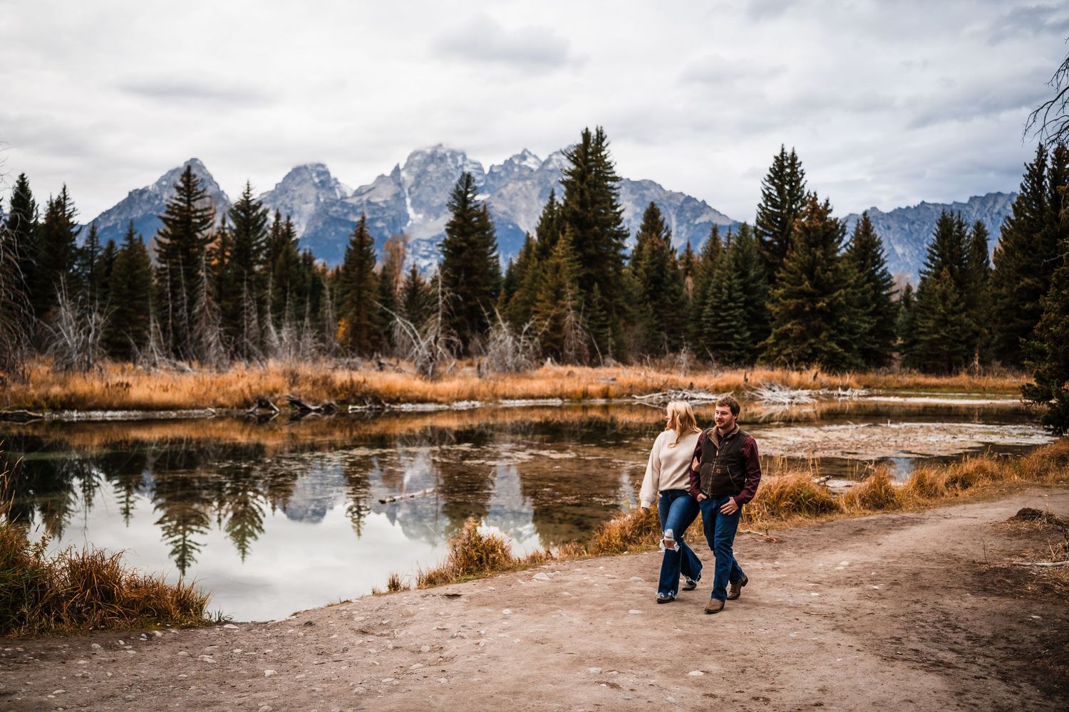 Panoramic view of snow-capped mountains with evergreen trees and a winding path along a tranquil lake in Grand Teton National Park.