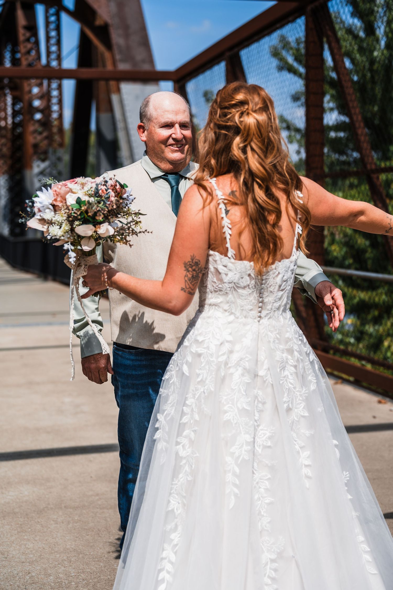 A bride in a white lace gown walks along an industrial bridge with wedding party members.