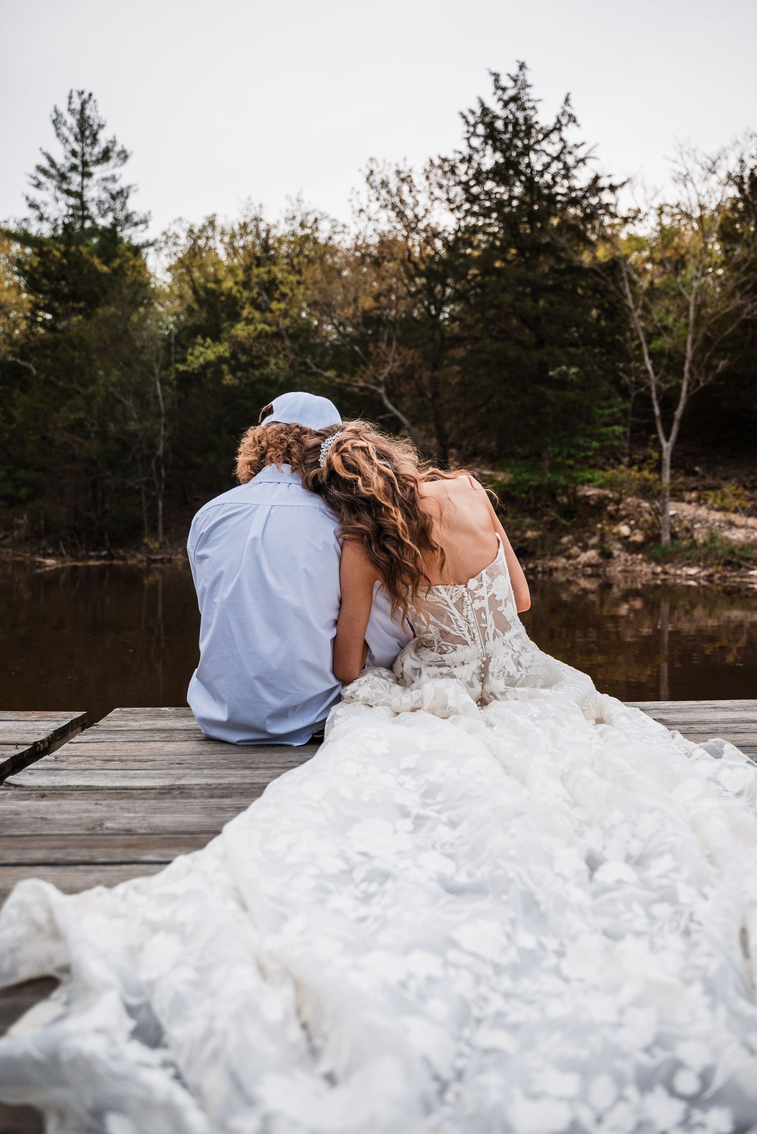 A couple sits together on a dock overlooking a lake with the bride wearing a flowing white wedding dress.