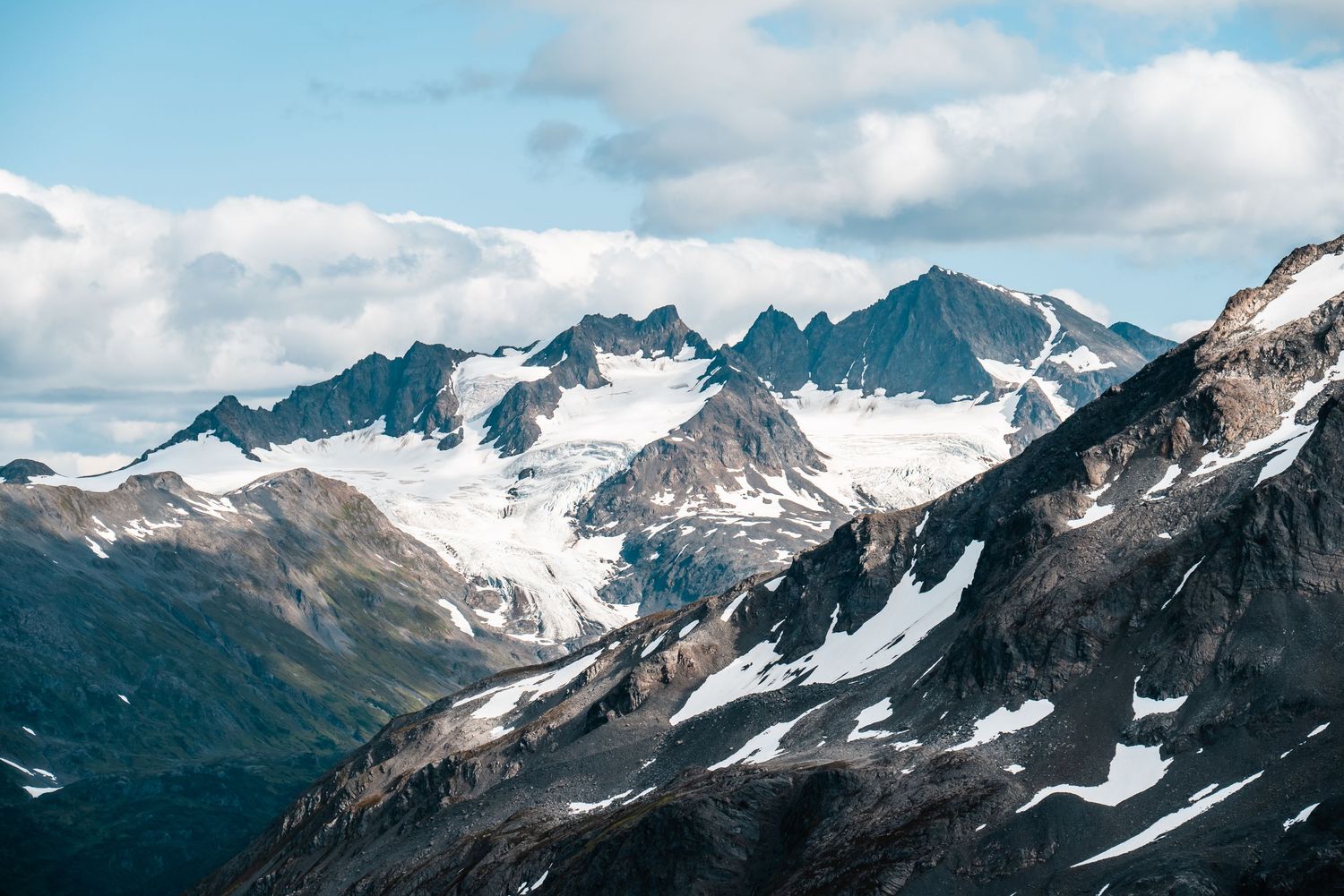 Dramatic mountain peaks covered in snow and glaciers rise against a cloudy sky in a rugged alpine landscape.
