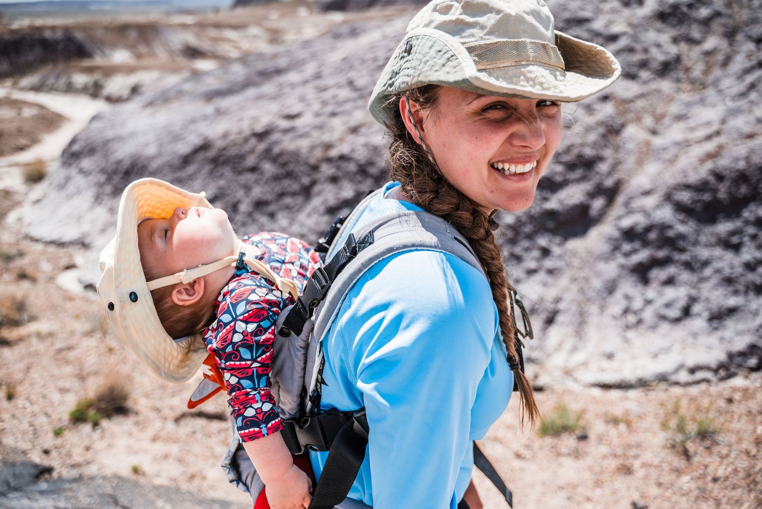 A hiker wearing a blue shirt and baseball cap carries a baby in a colorful backpack carrier while exploring outdoors.