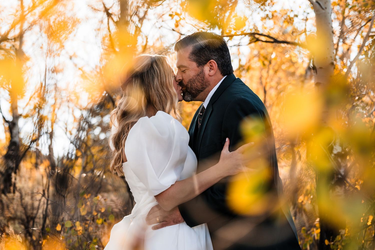 A couple shares a romantic kiss in a sunlit autumn forest with golden leaves and warm lighting.