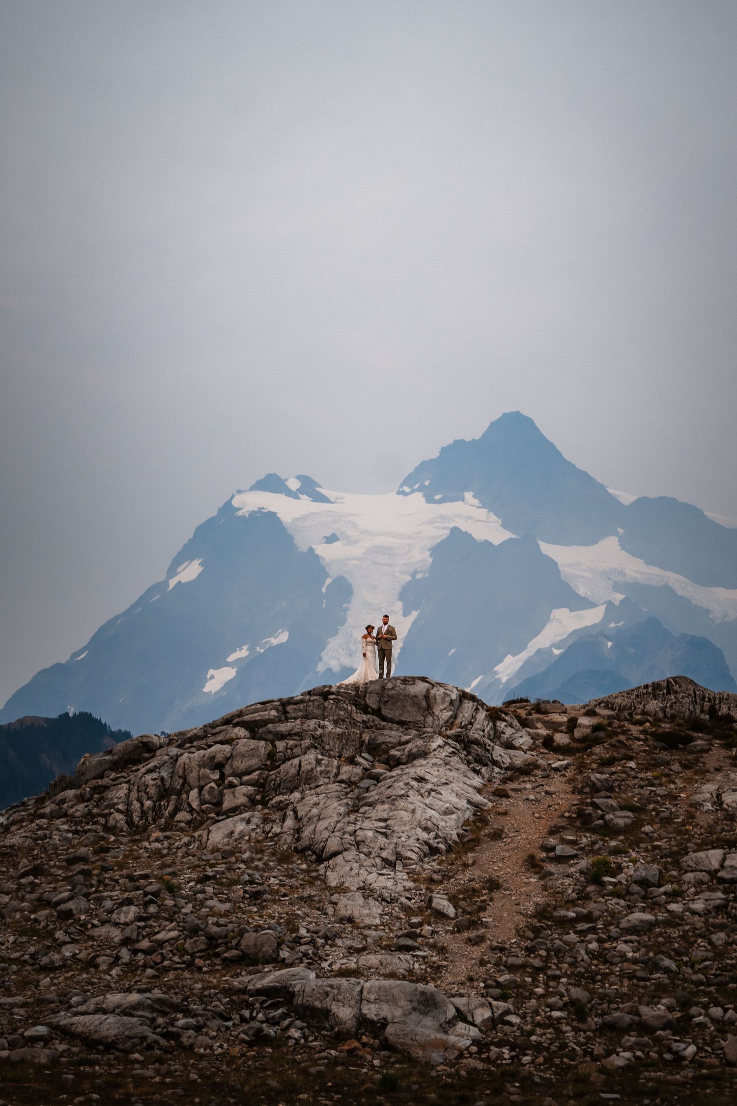Silhouettes stand atop rocky mountain ridge with snow-capped peaks and glaciers in the background.