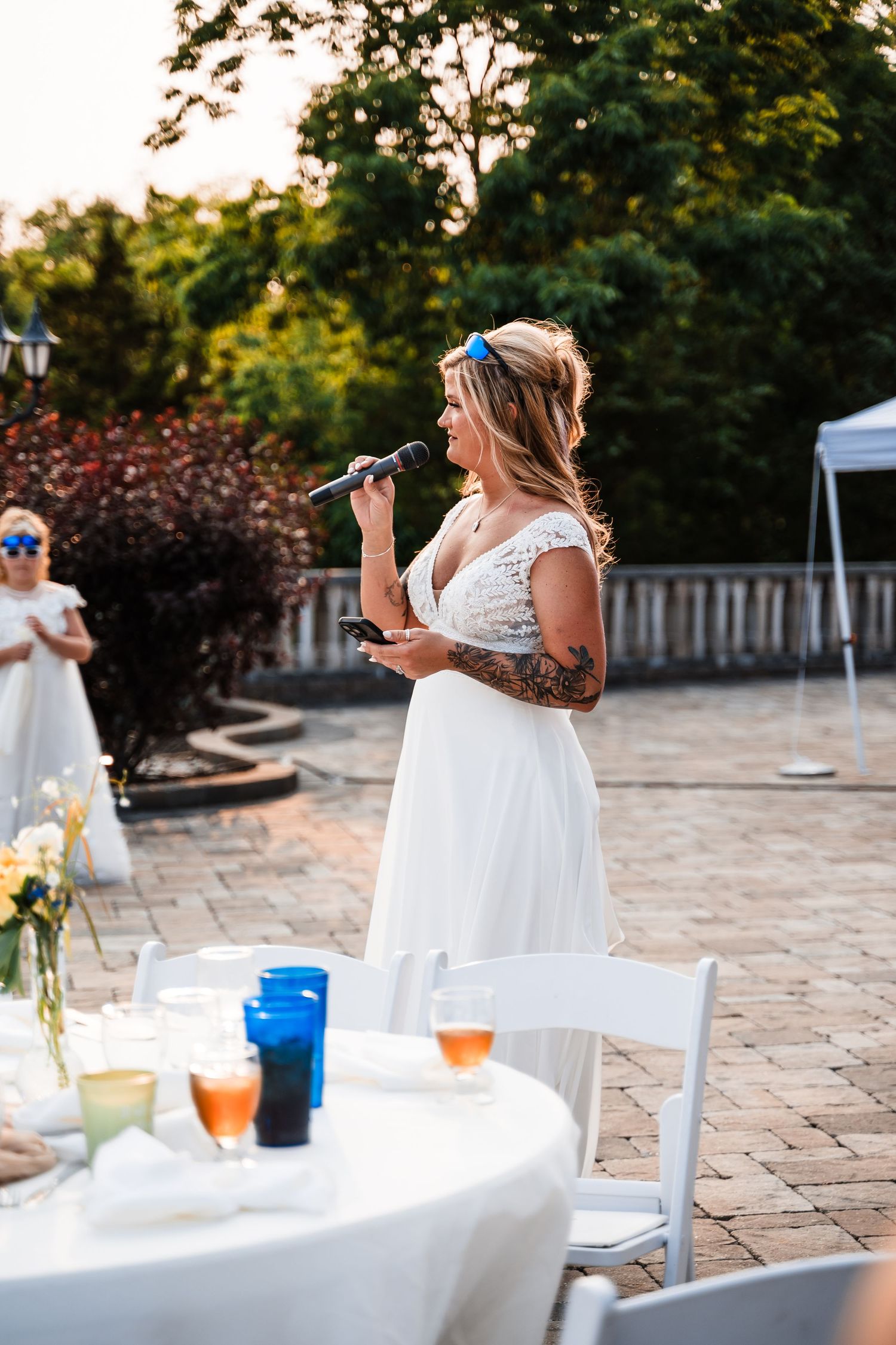 Person in white dress making a speech at an outdoor wedding reception with drinks on tables.