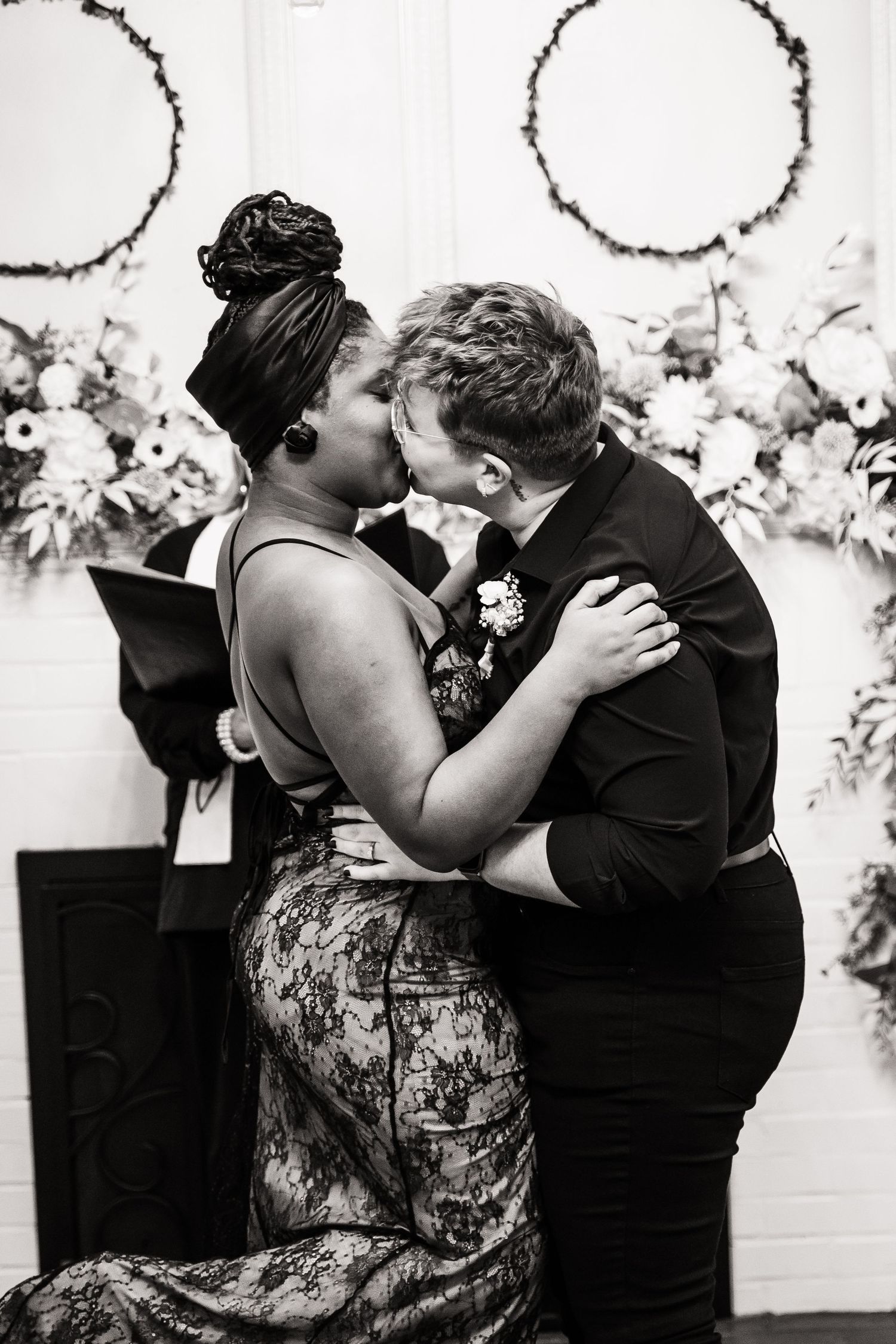 A romantic black and white photograph of a couple sharing their first kiss at a wedding ceremony with floral decorations.
