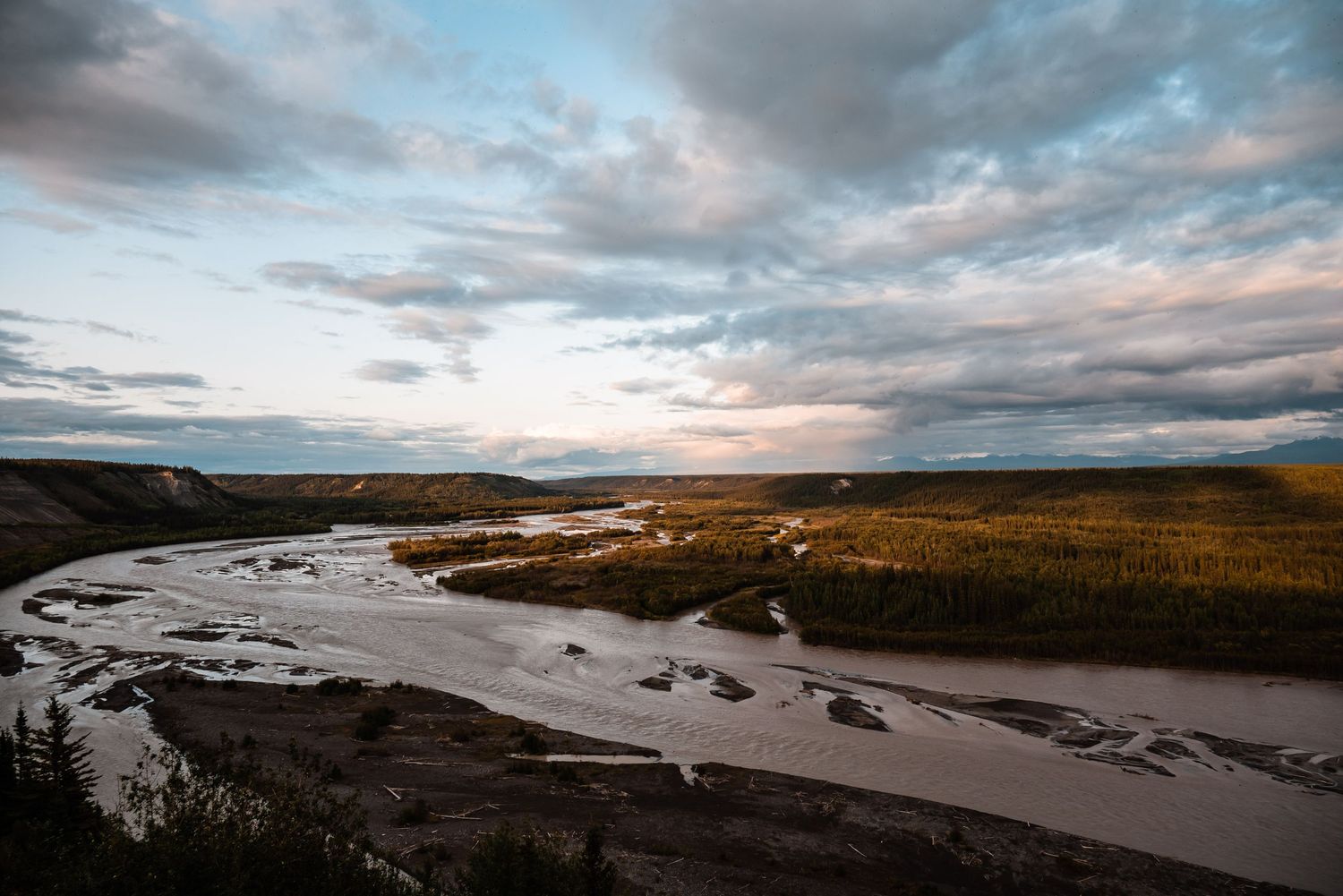 A winding river cuts through golden grasslands under dramatic sunset clouds in a remote landscape.