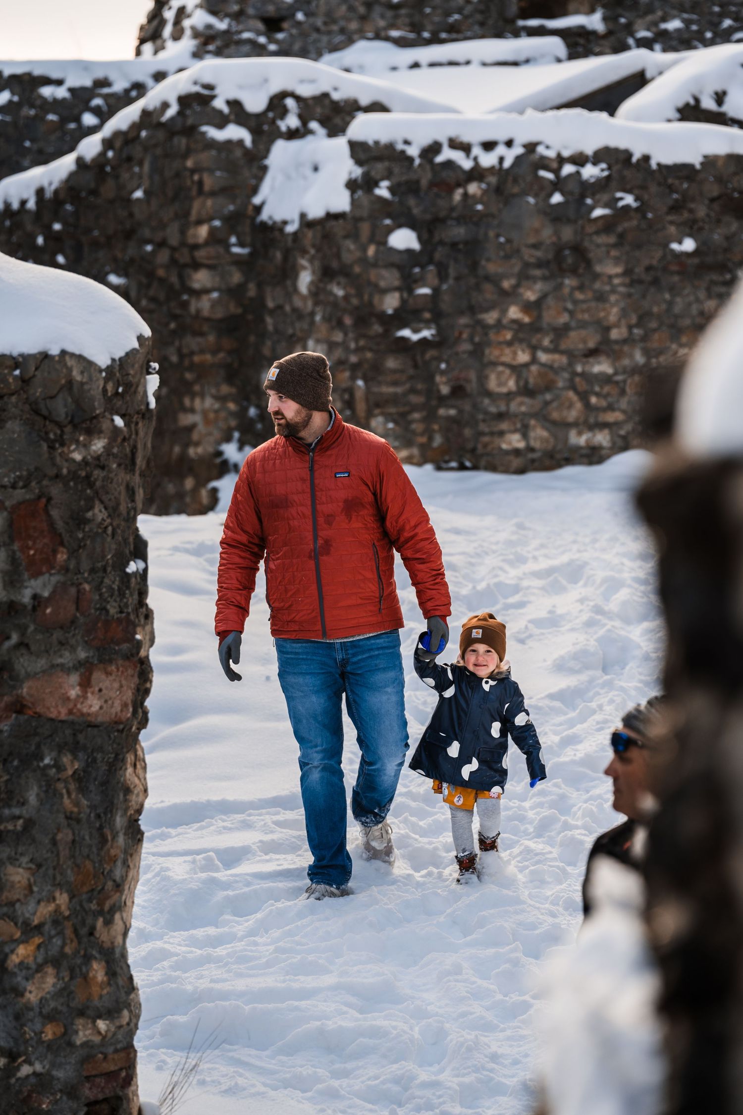 A pair walking through snowy stone ruins on a winter day wearing red and blue winter coats.