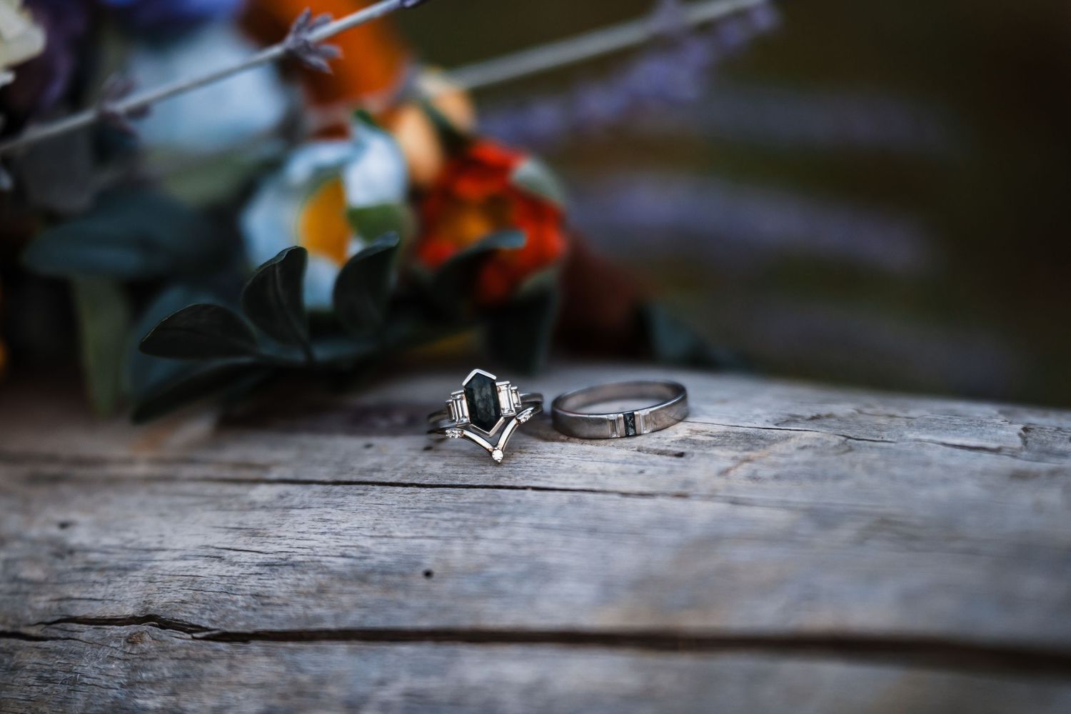 Silver engagement ring with dark gemstone on rustic wooden surface with colorful floral bouquet in background.