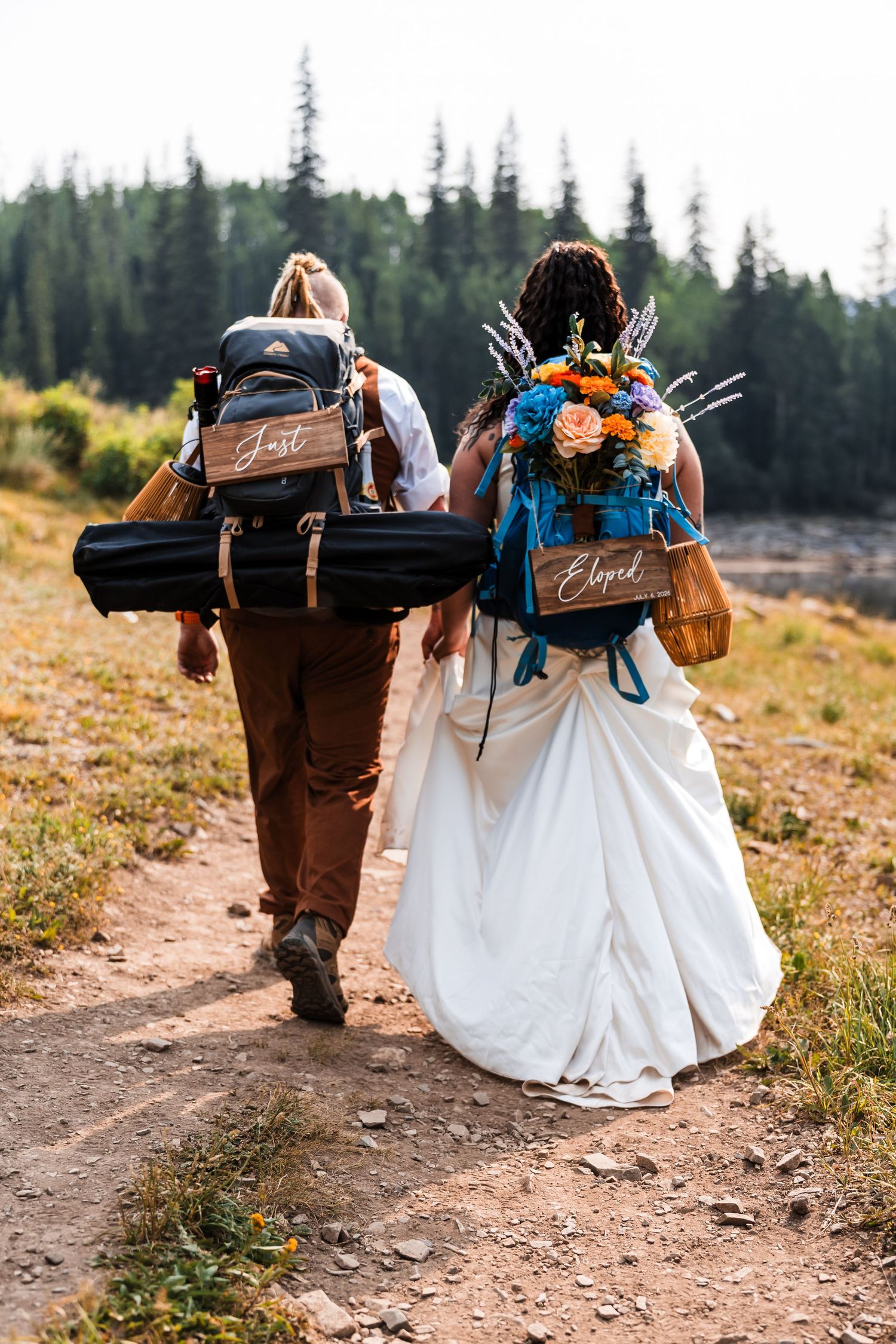 A bride and groom in wedding attire hike a mountain trail carrying backpacks labeled Just Eloped.