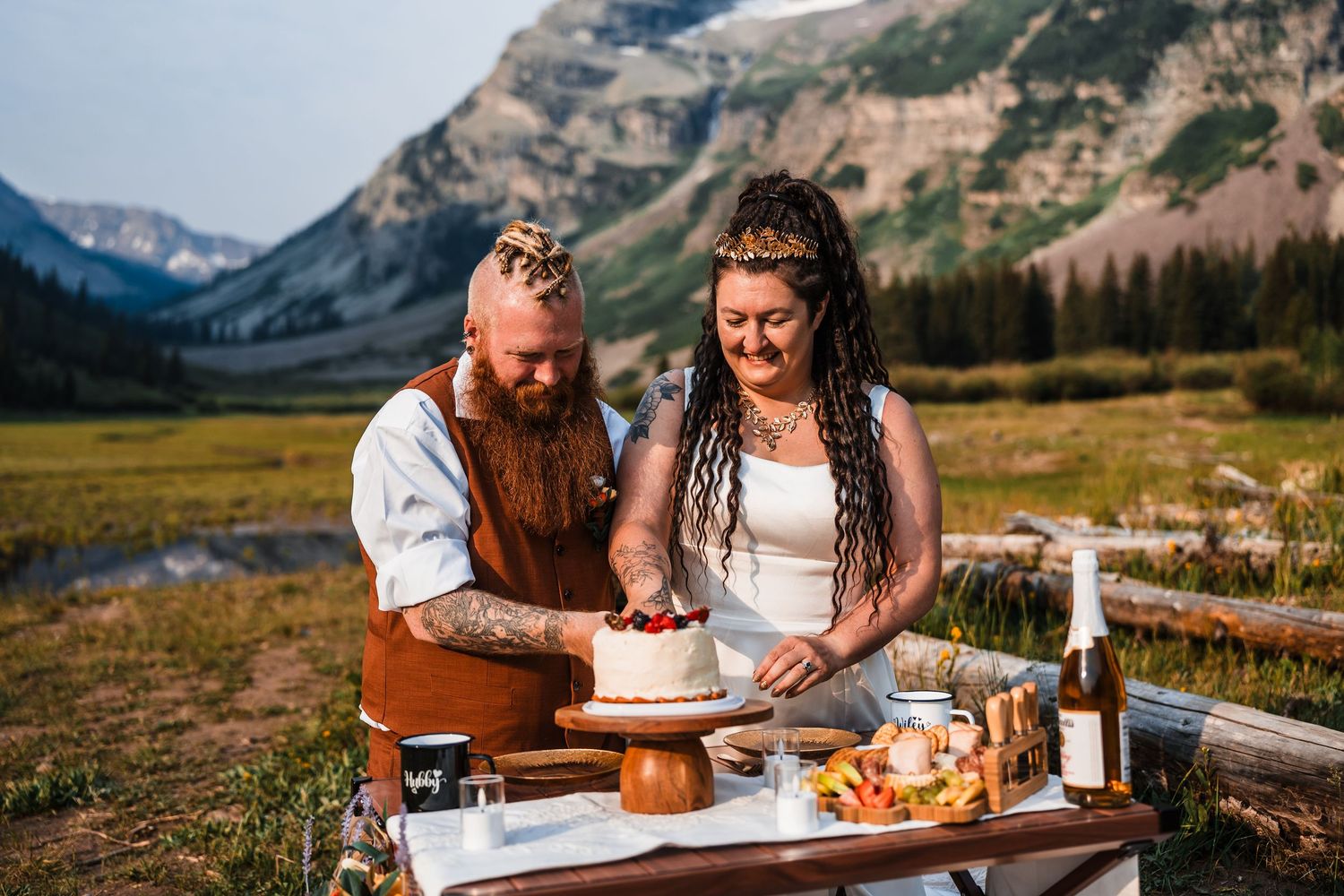 Couple cuts white wedding cake together at outdoor table with stunning mountain meadow backdrop during elopement celebration.